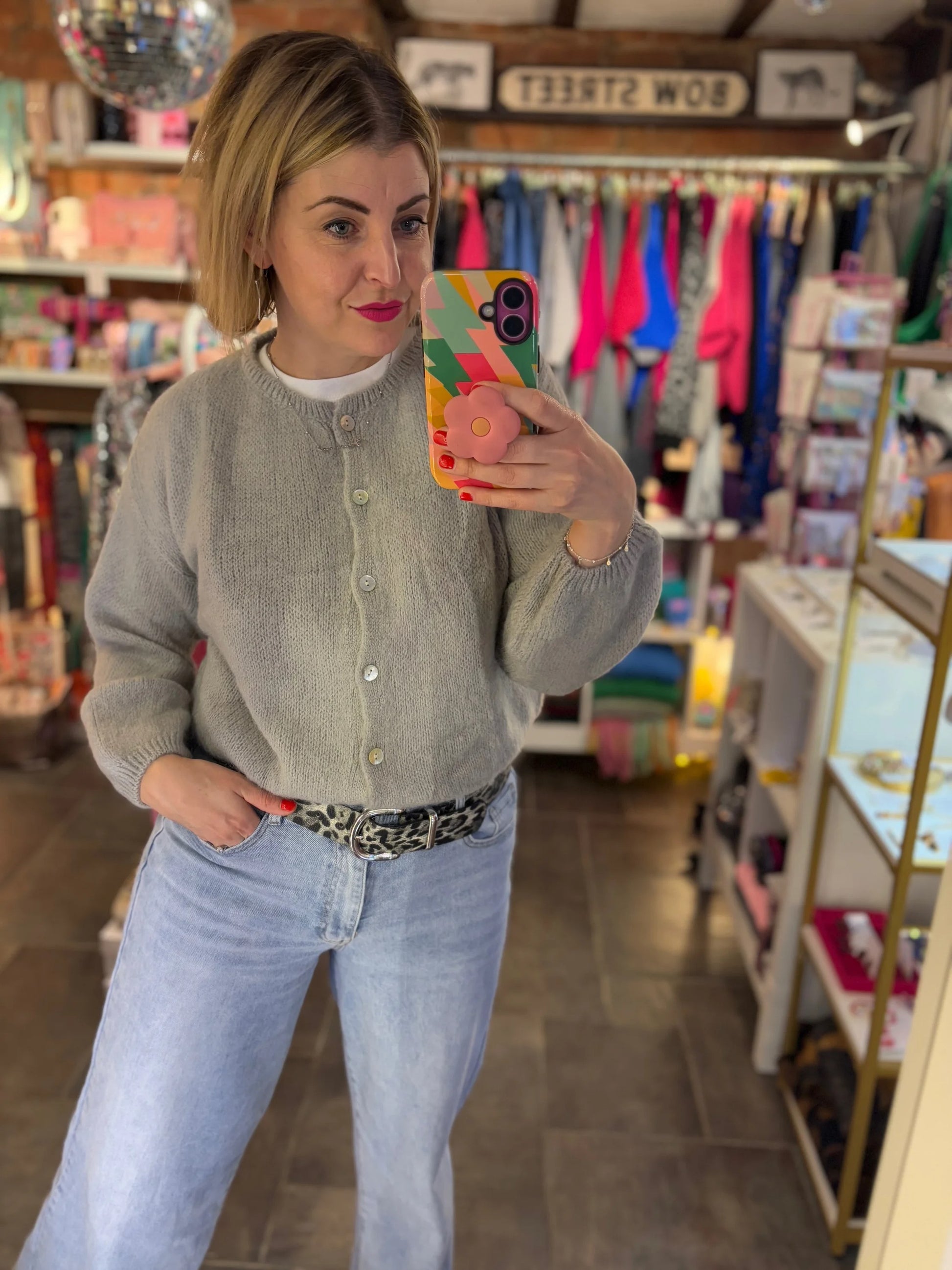 Woman taking a mirror selfie in a store wearing a gray cardigan and light blue jeans.