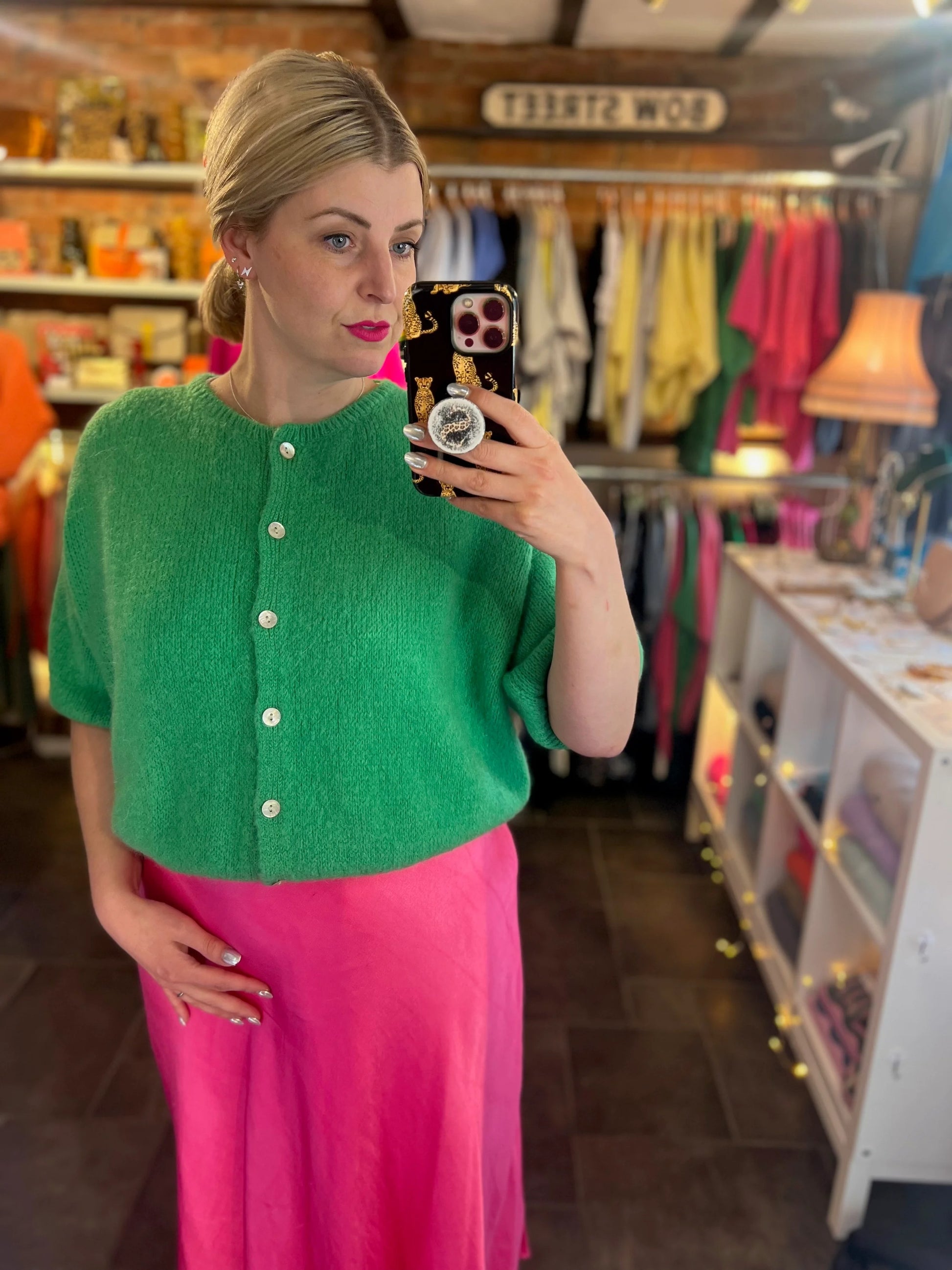 Woman taking a mirror selfie wearing a green cardigan and pink skirt in a store.