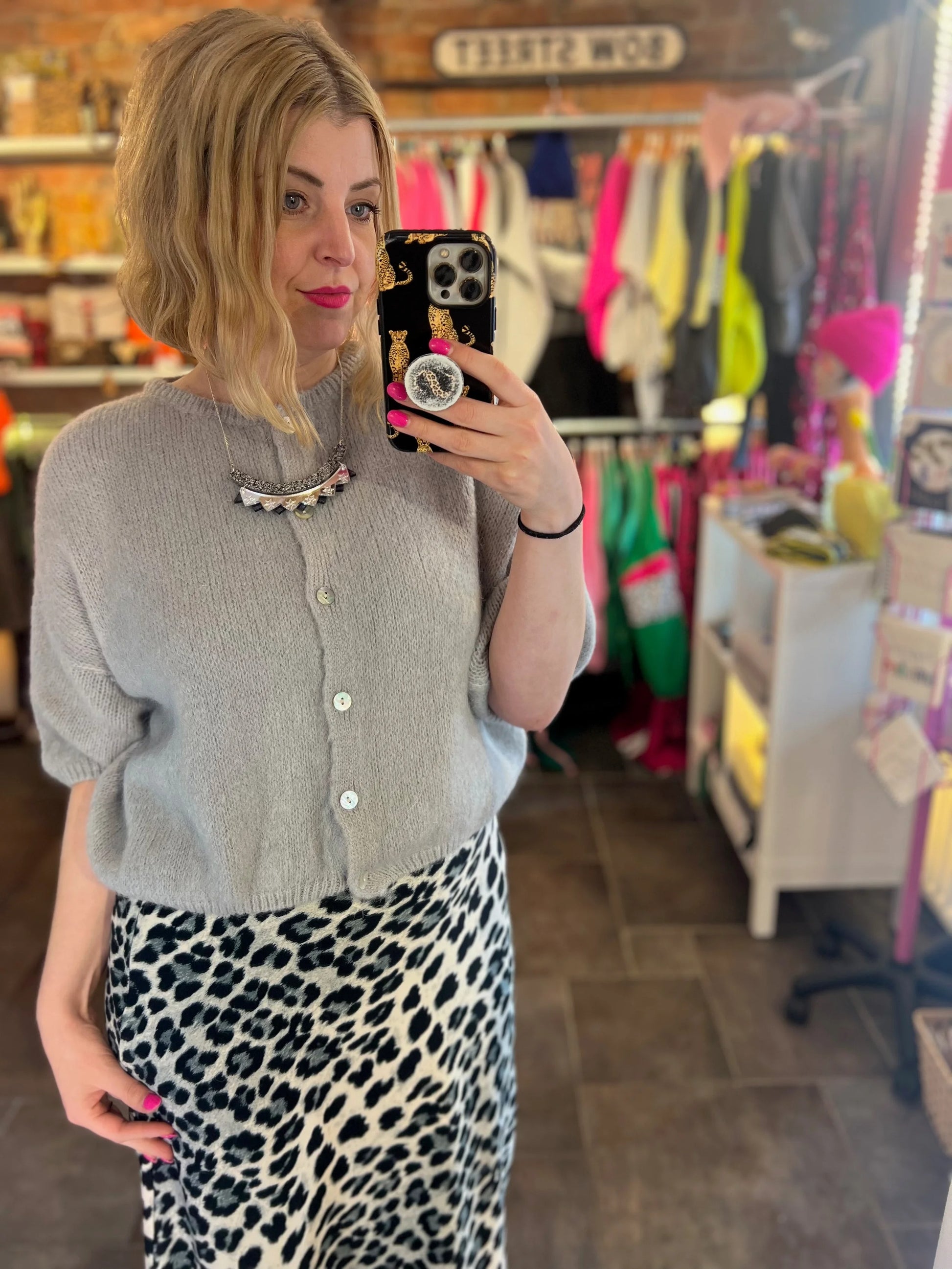 Person taking a mirror selfie wearing a gray cardigan and leopard print skirt in a store.