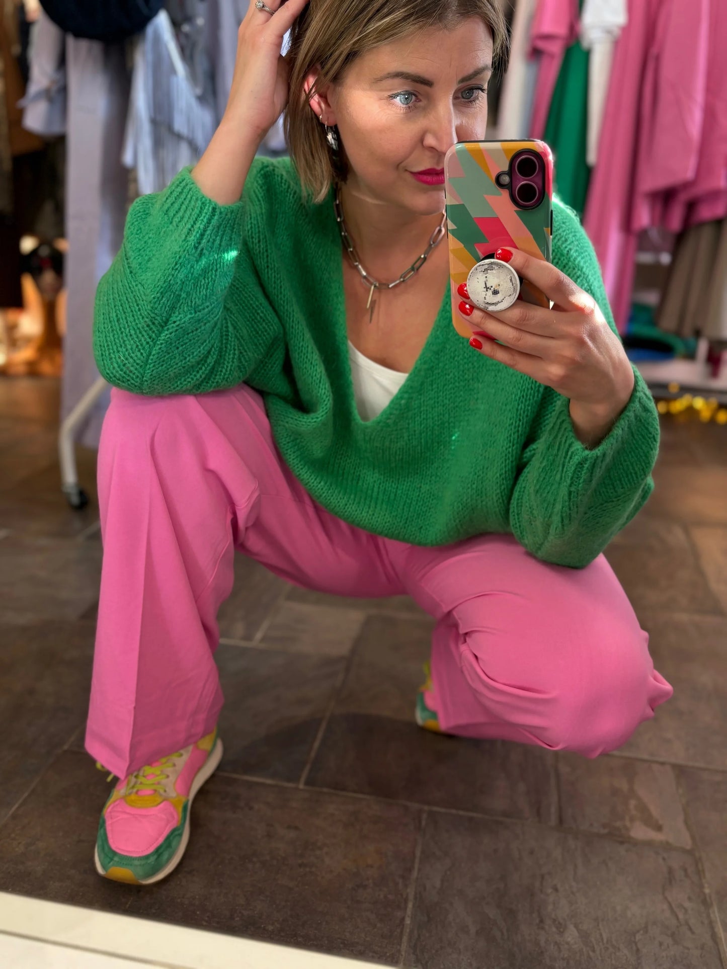 Person wearing a green sweater and pink pants taking a mirror selfie in a store.