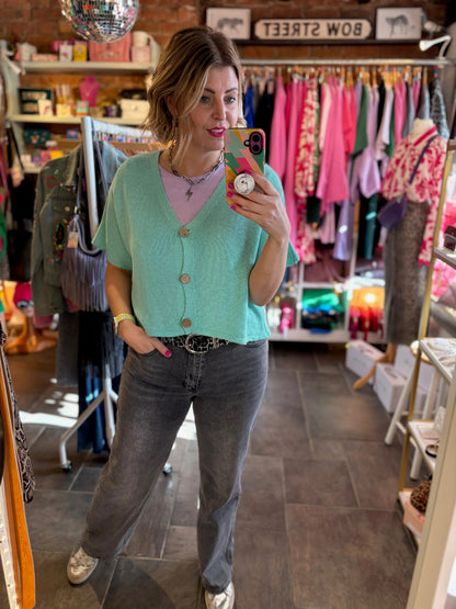 Woman taking a mirror selfie in a clothing store wearing a green cardigan and gray jeans.