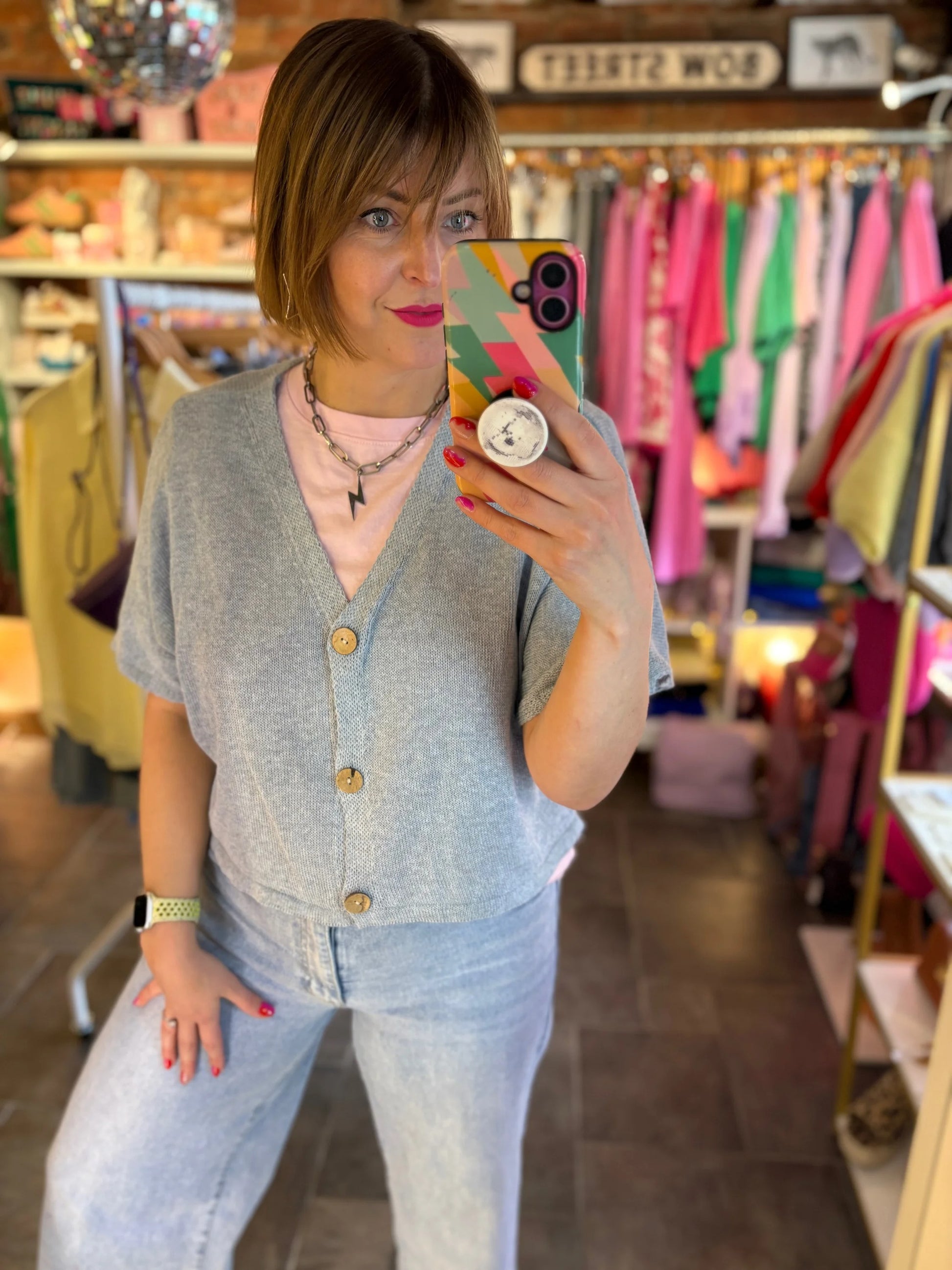Person taking a mirror selfie in a clothing store wearing a gray cardigan and light blue jeans.