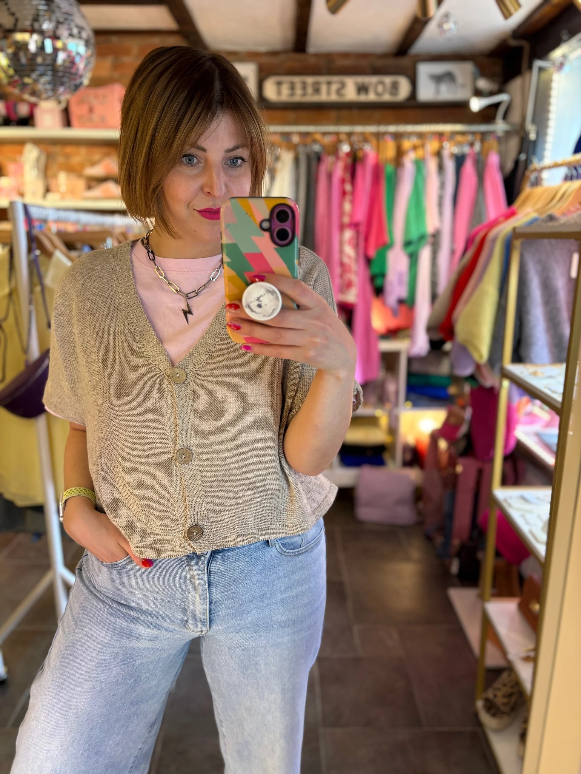 Person taking a mirror selfie in a clothing store with colorful shirts on racks.