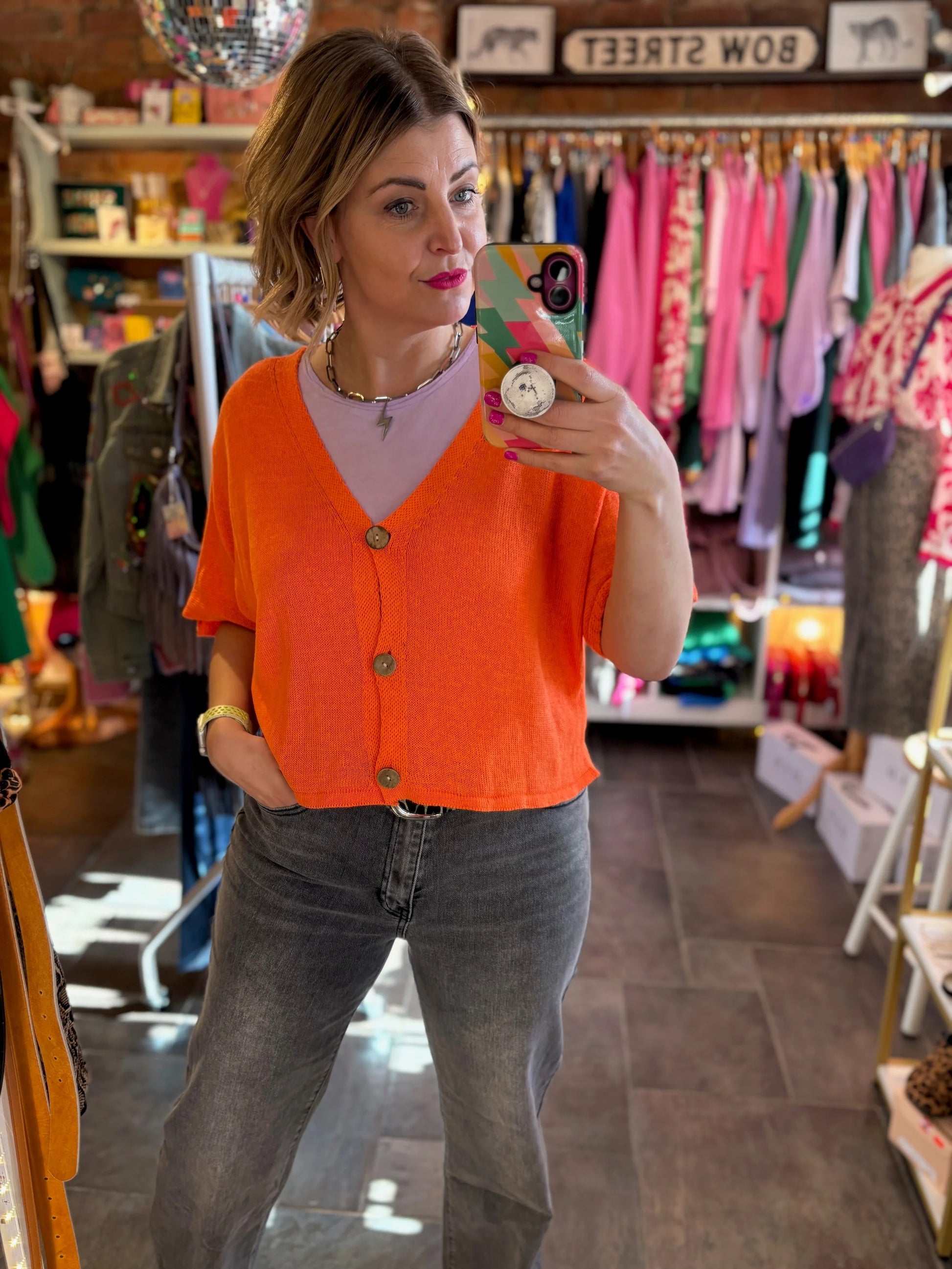 Woman taking a mirror selfie wearing an orange cardigan and gray jeans in a store.