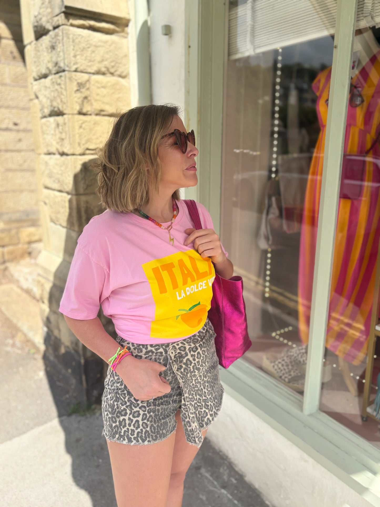 Woman wearing a pink shirt with a yellow 'ITALY' design, leopard print shorts, and sunglasses, standing outside a store.