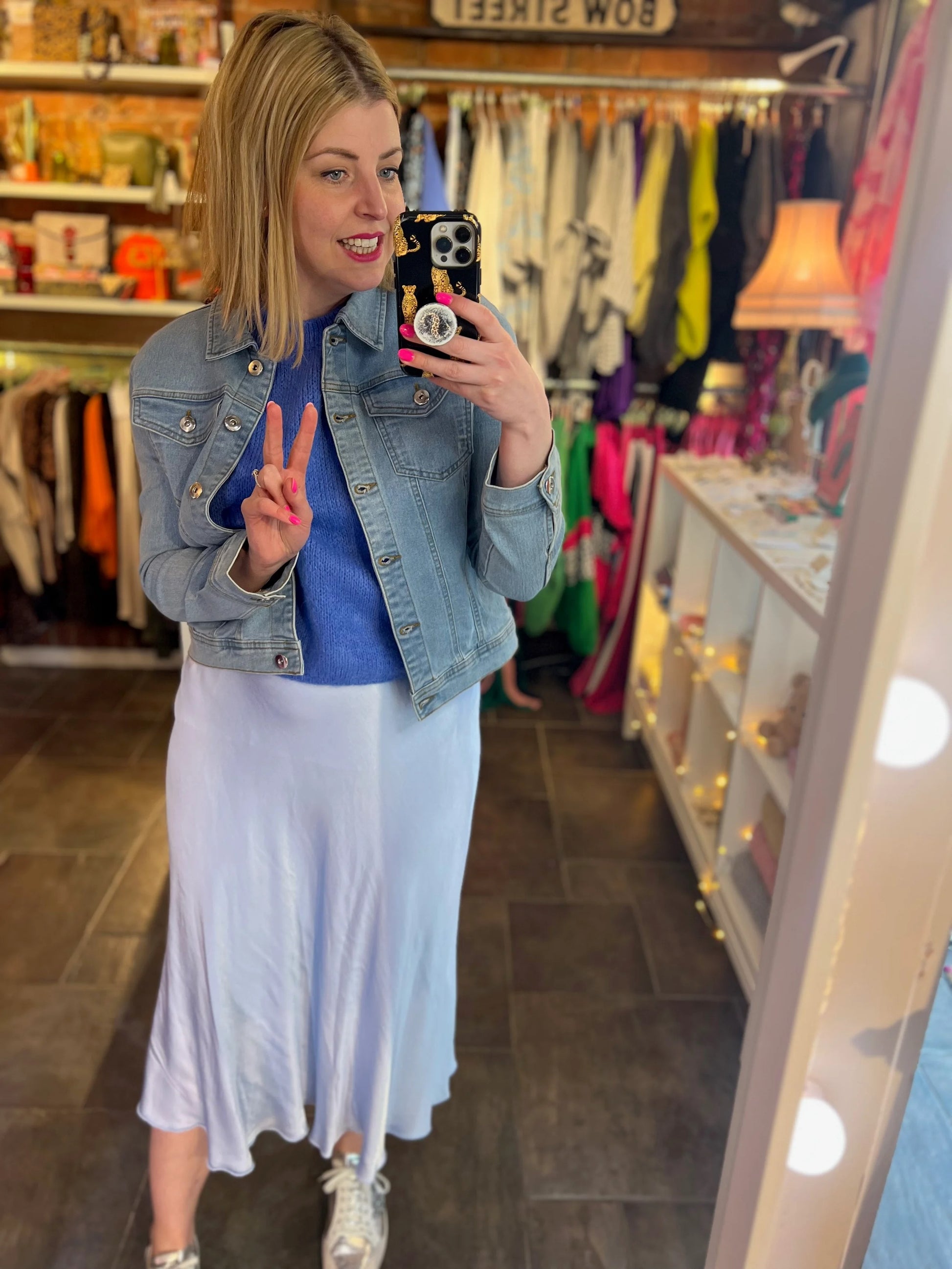 Woman taking a mirror selfie in a clothing store, wearing a denim jacket and white skirt.