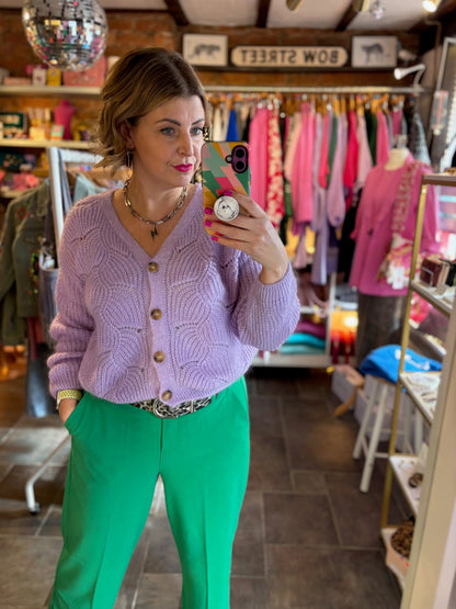 Woman taking a mirror selfie wearing a purple cardigan and green pants in a store.