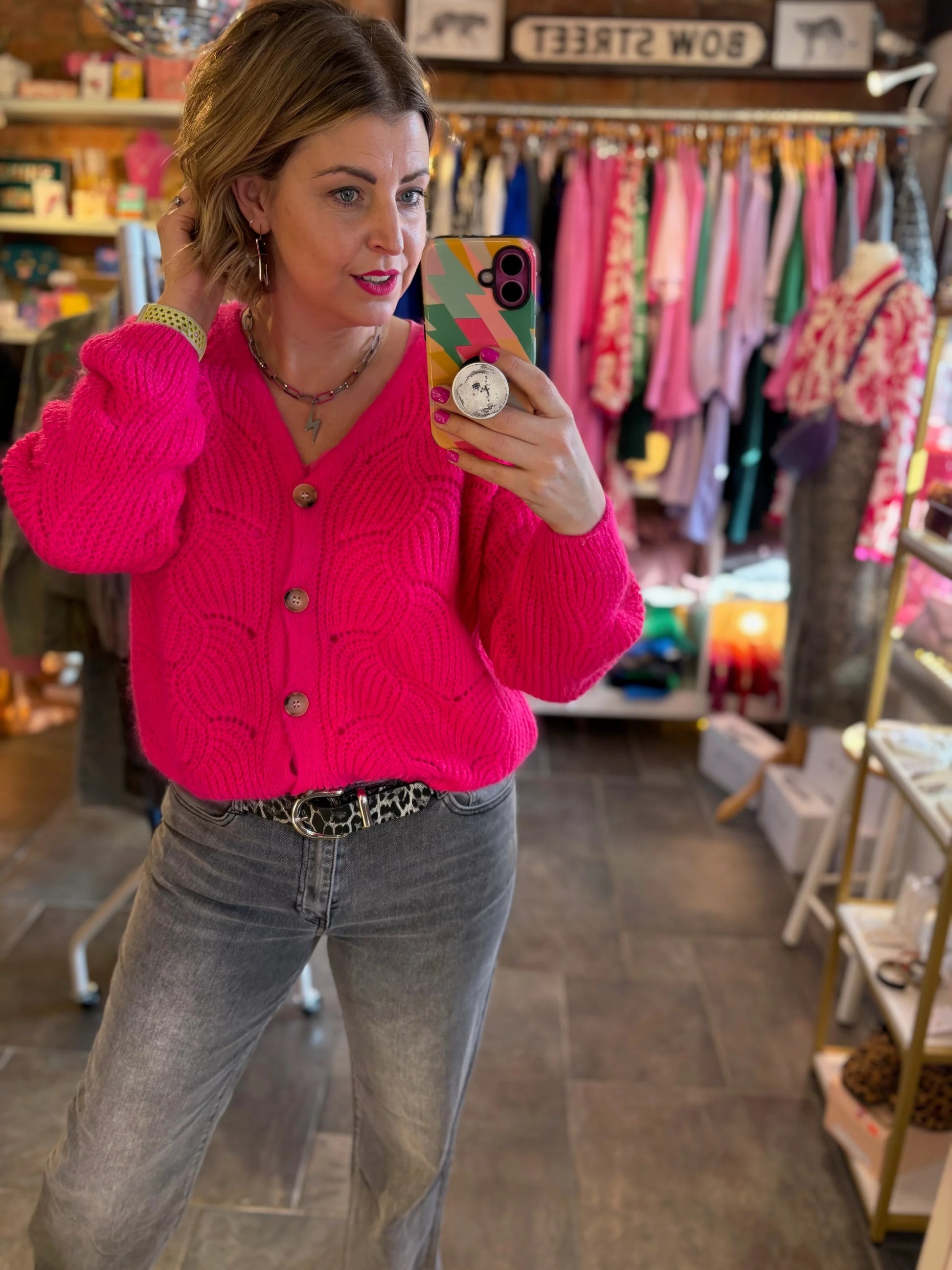 Woman taking a mirror selfie wearing a bright pink cardigan in a store.
