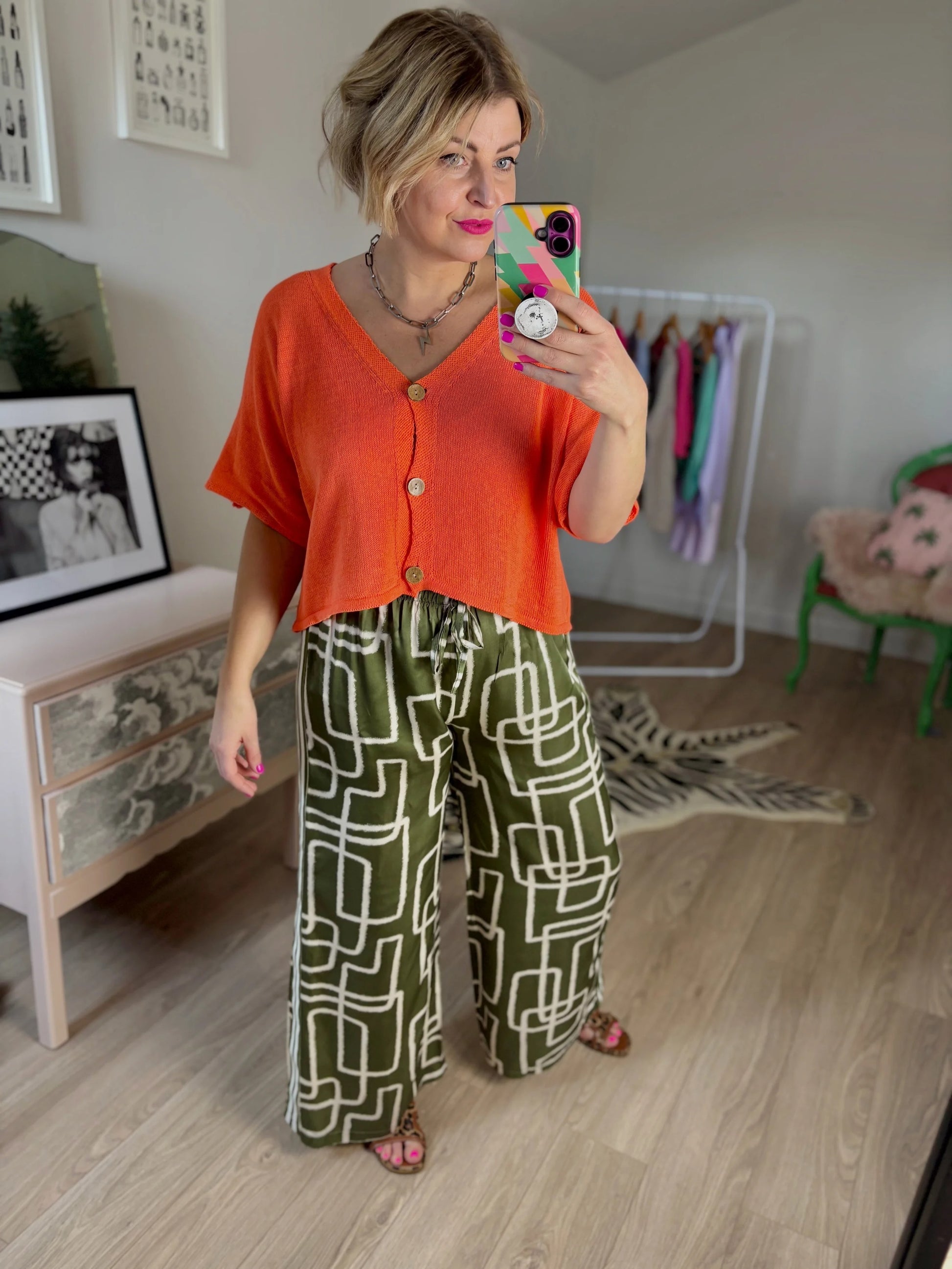 Woman taking a mirror selfie wearing an orange top and green patterned pants in a room.