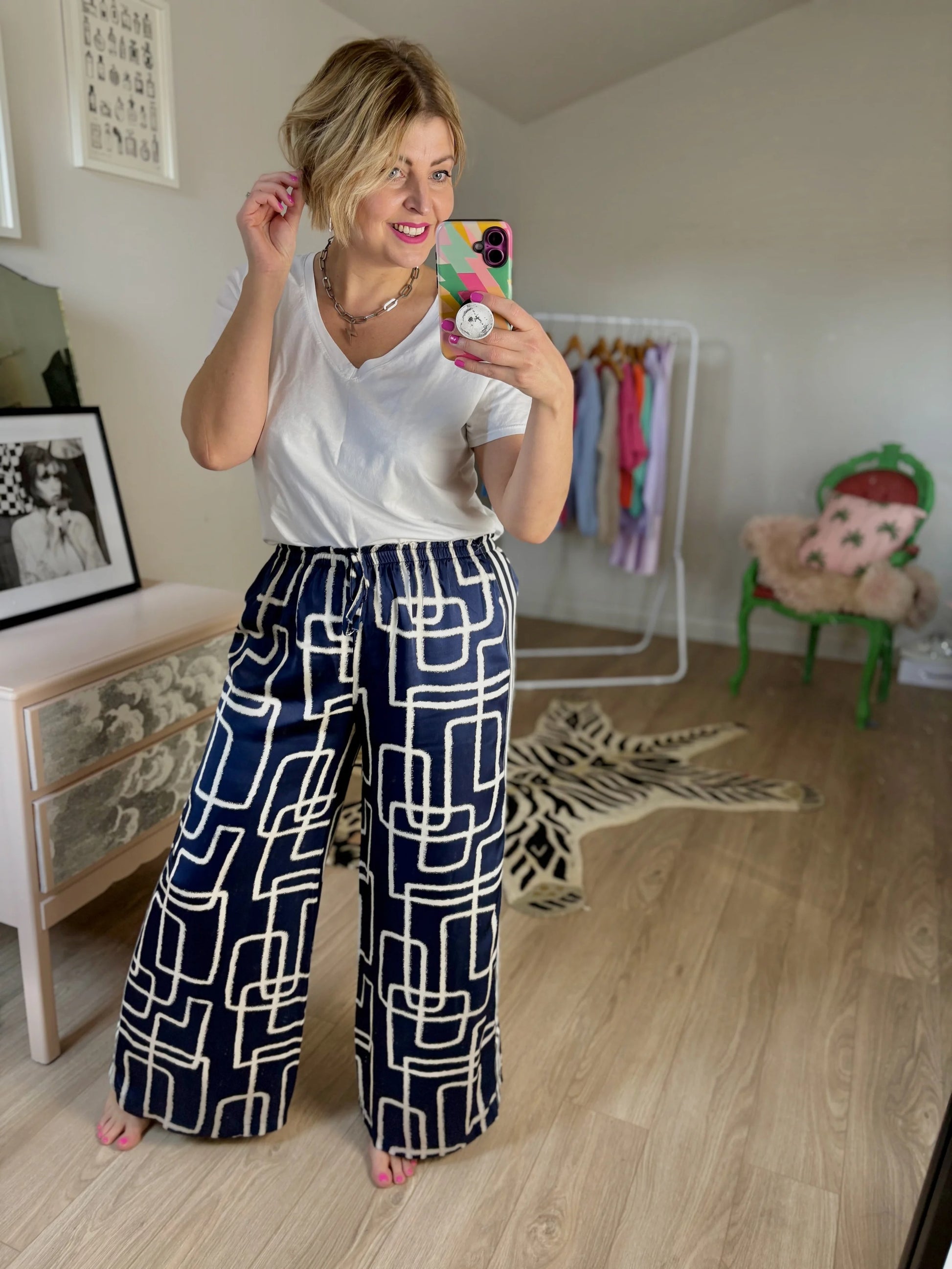 Woman taking a mirror selfie wearing a white top and navy patterned pants in a room.