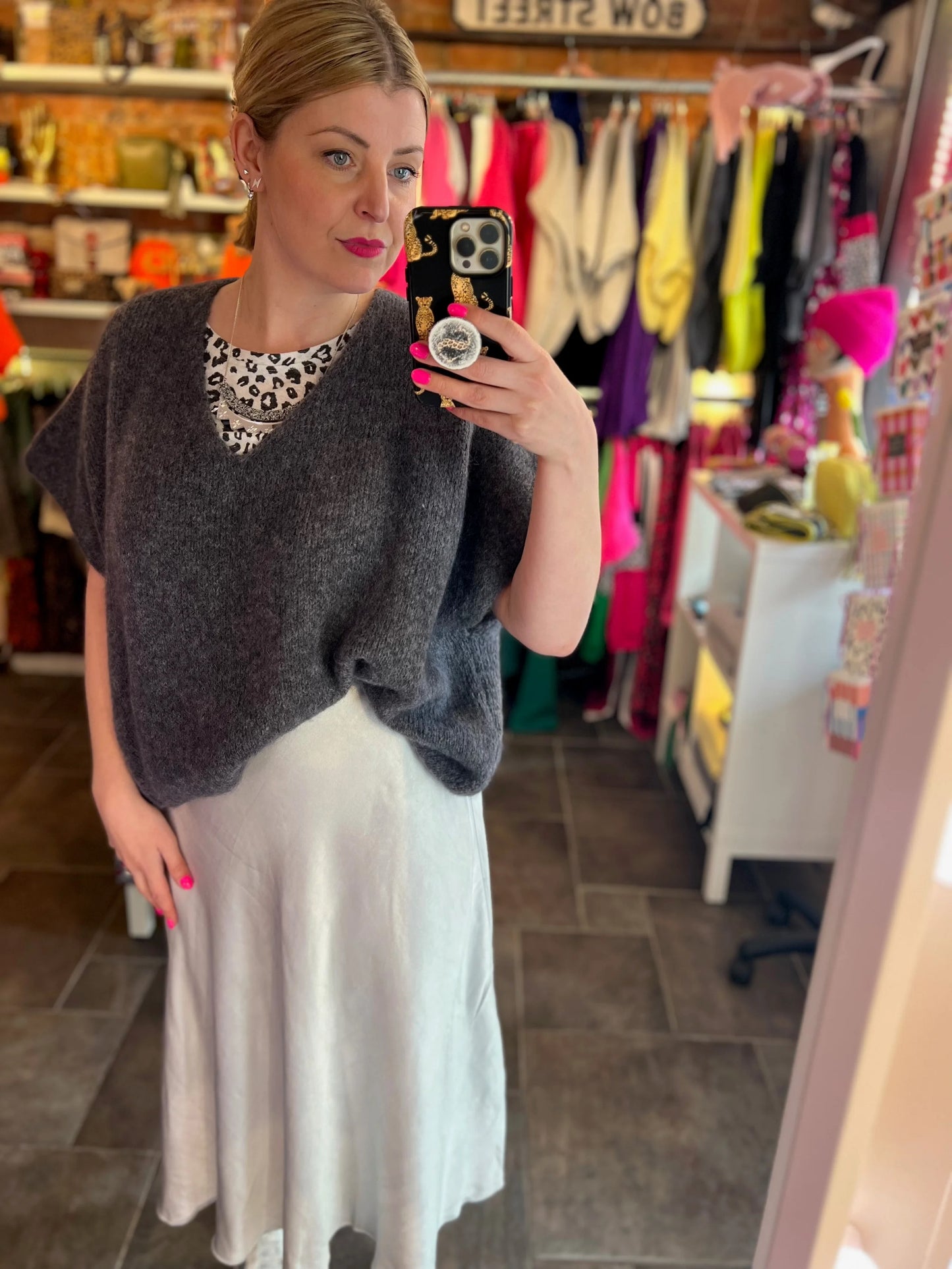 Woman taking a mirror selfie wearing a gray sweater and white skirt in a store.