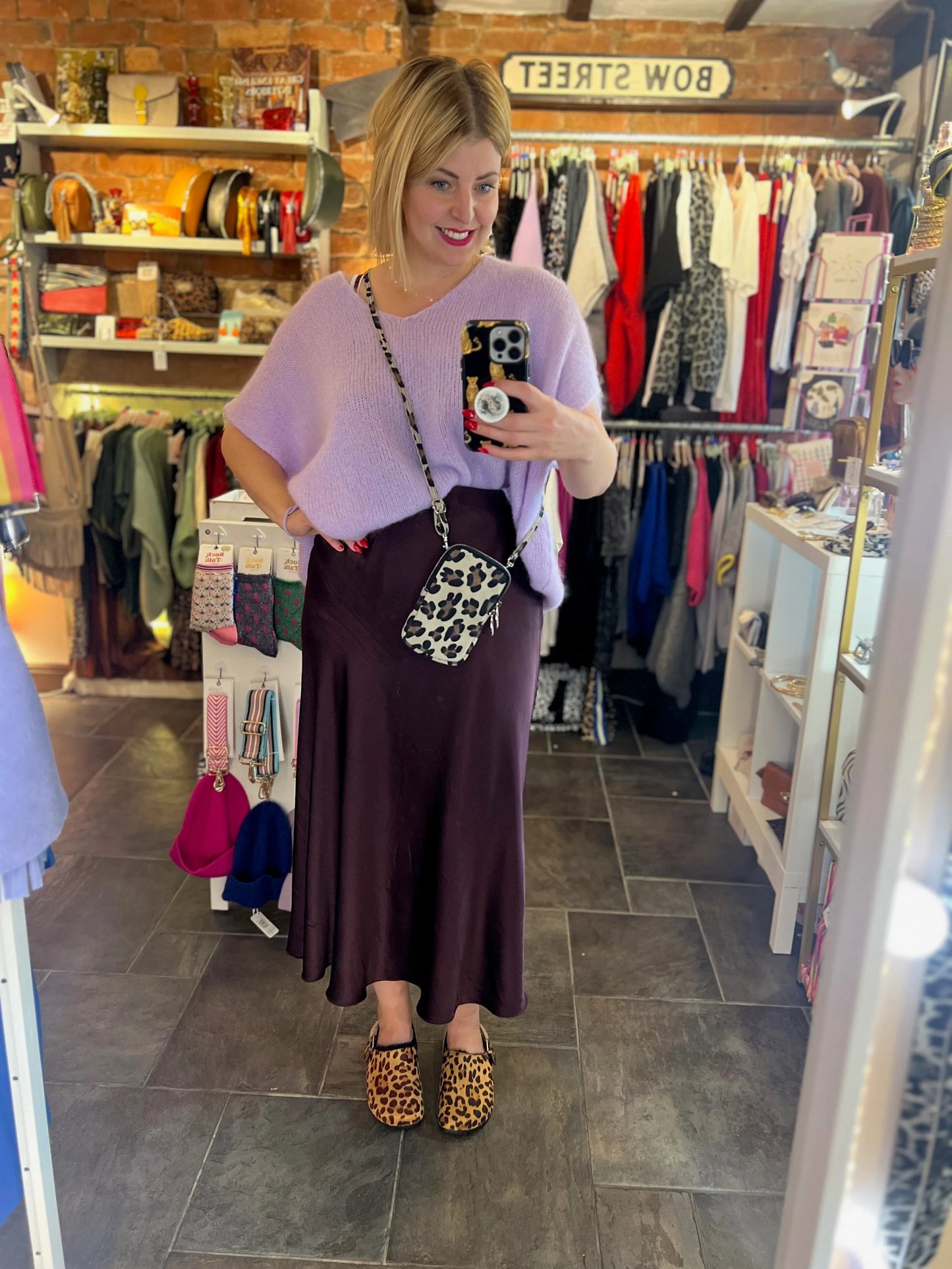 Woman taking a mirror selfie in a clothing store wearing a purple top and brown skirt.