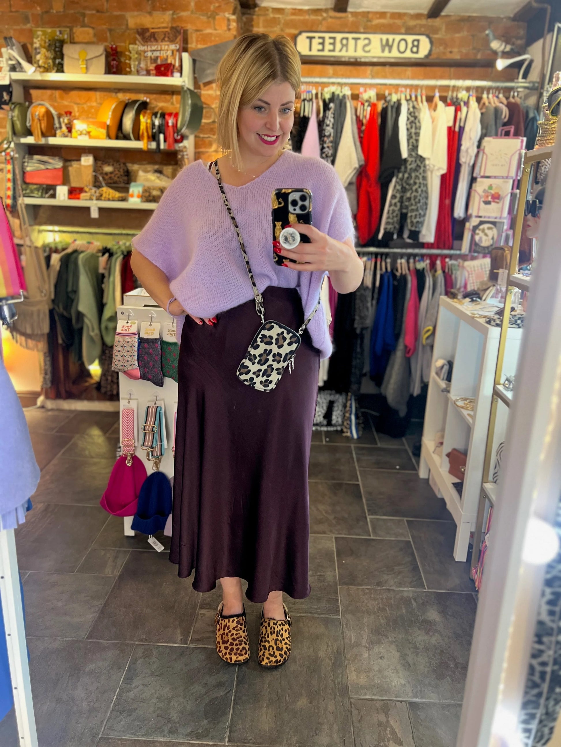 Woman taking a mirror selfie in a clothing store wearing a purple top and brown skirt.