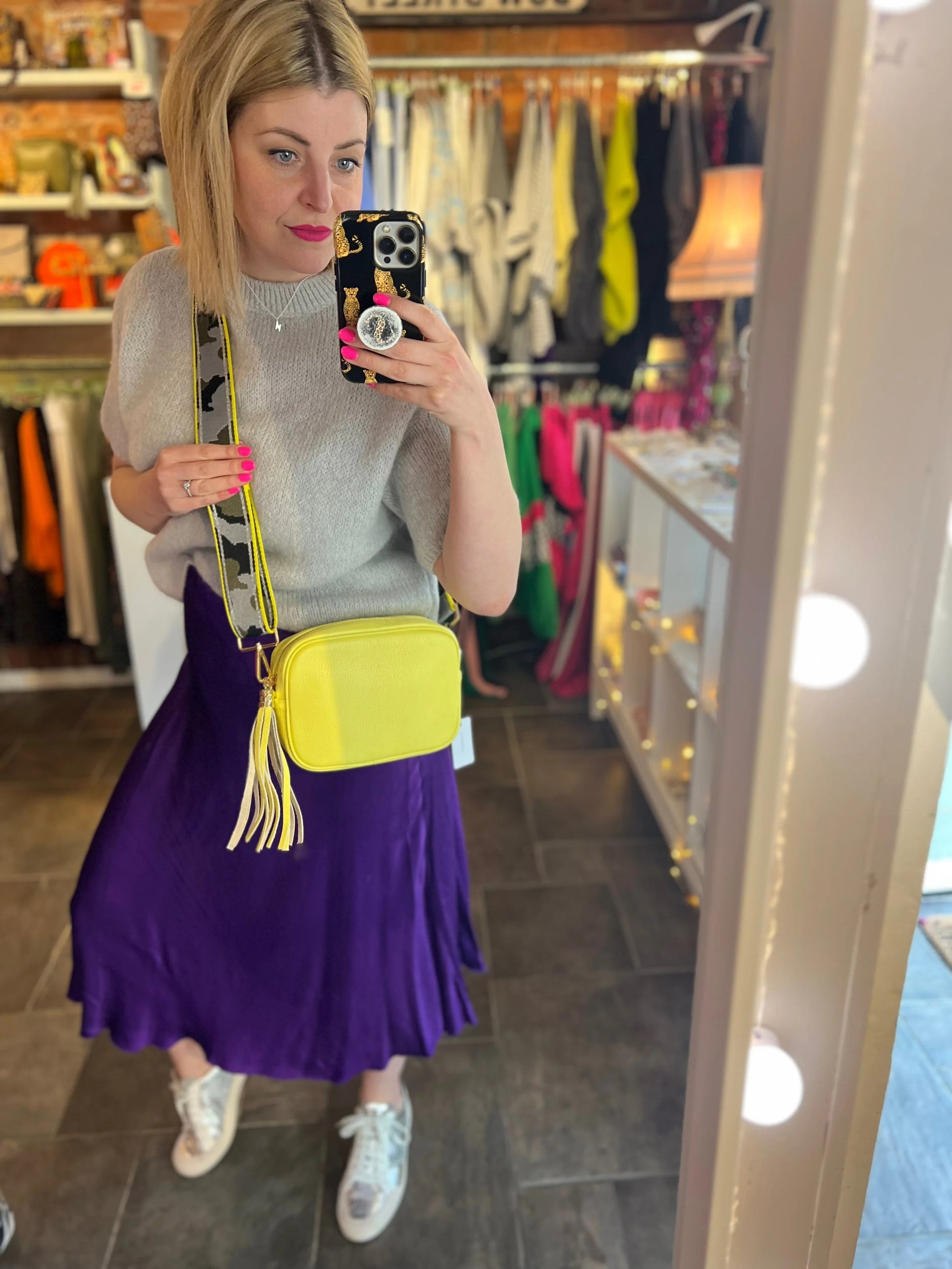 Woman taking a mirror selfie wearing a purple skirt and yellow bag in a store.