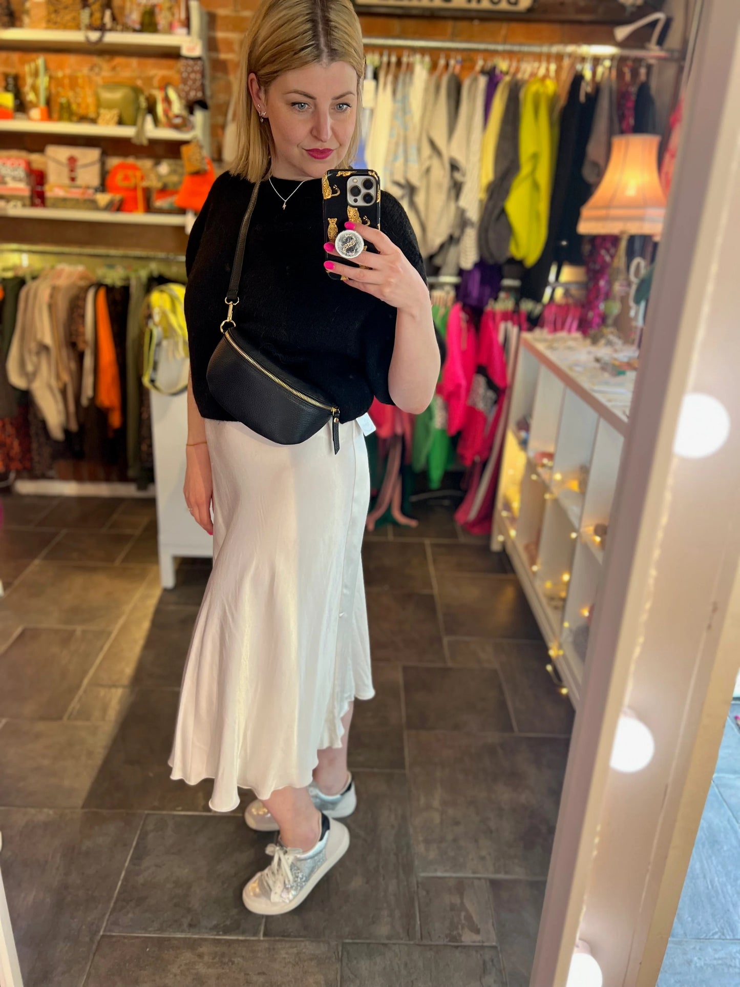 Woman taking a mirror selfie in a clothing store wearing a black top, white skirt, and sneakers.