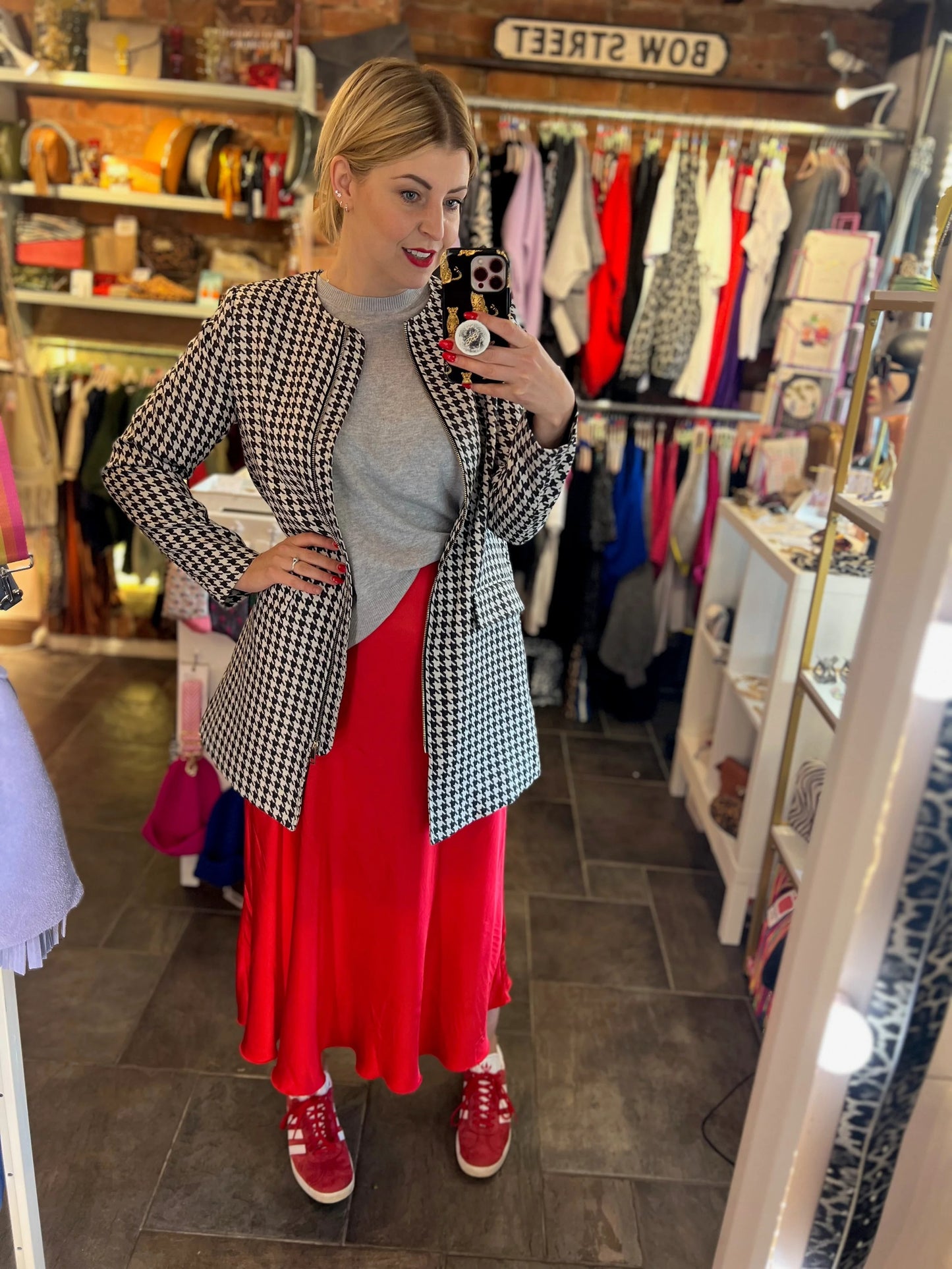 Woman taking a mirror selfie in a clothing store wearing a checkered jacket, gray turtleneck, red skirt, and red shoes.