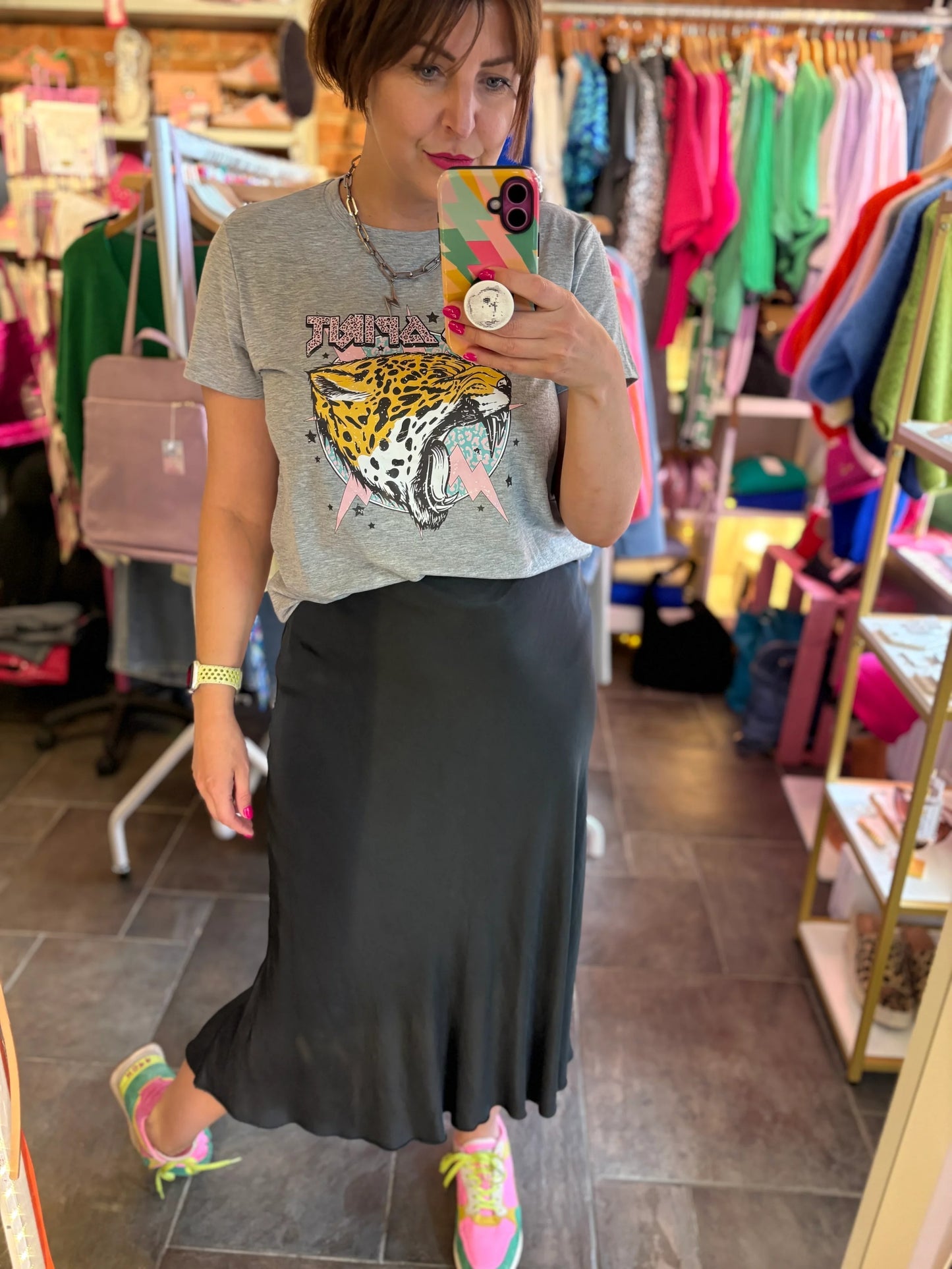 Woman taking a mirror selfie in a clothing store wearing a graphic t-shirt and black skirt.