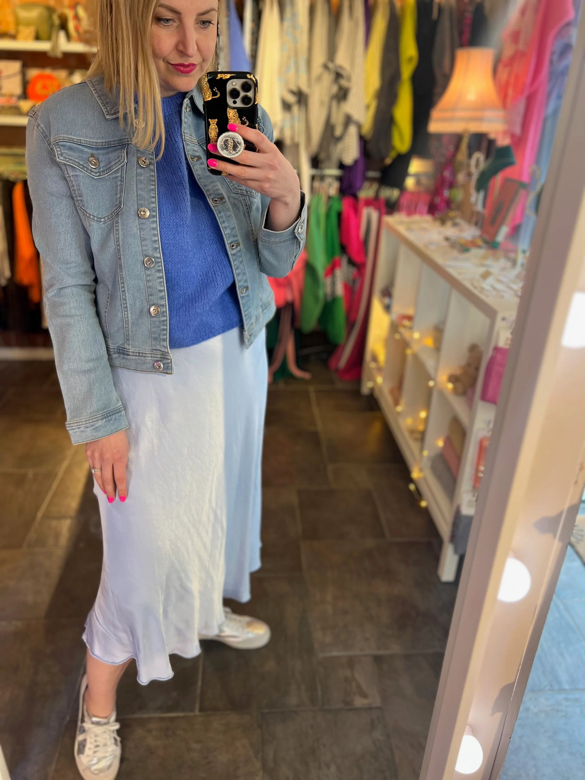 Woman taking a mirror selfie in a clothing store wearing a denim jacket, blue top, and white skirt.