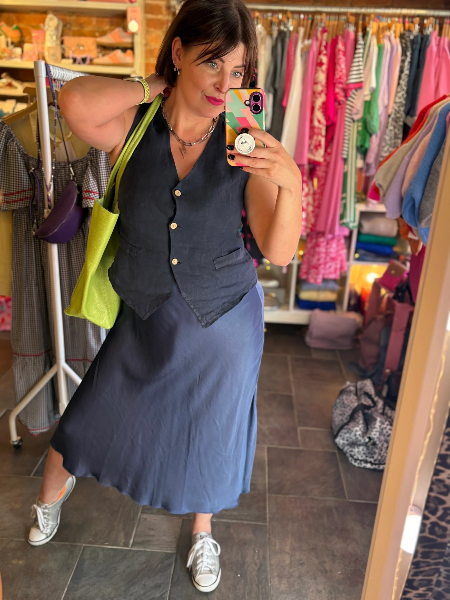 Woman taking a mirror selfie wearing a blue dress and black vest in a clothing store.