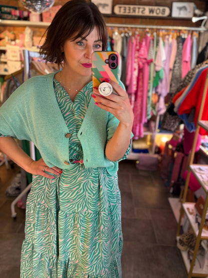 Woman taking a mirror selfie wearing a green dress with a patterned phone case in a store.
