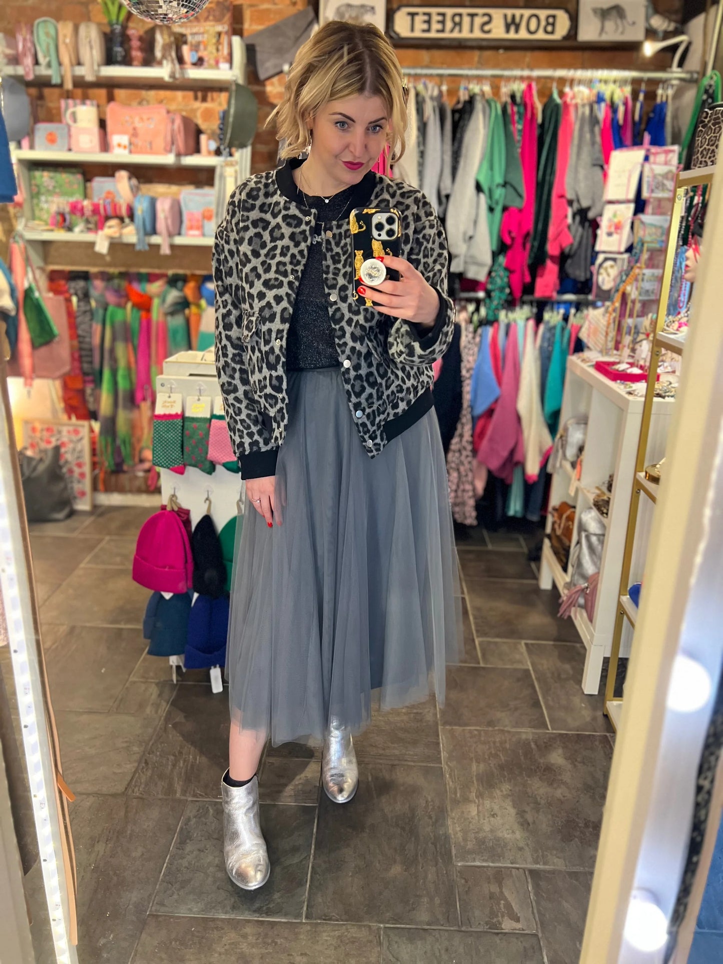 Woman taking a mirror selfie in a clothing store wearing a leopard print jacket, gray tulle skirt, and silver shoes.