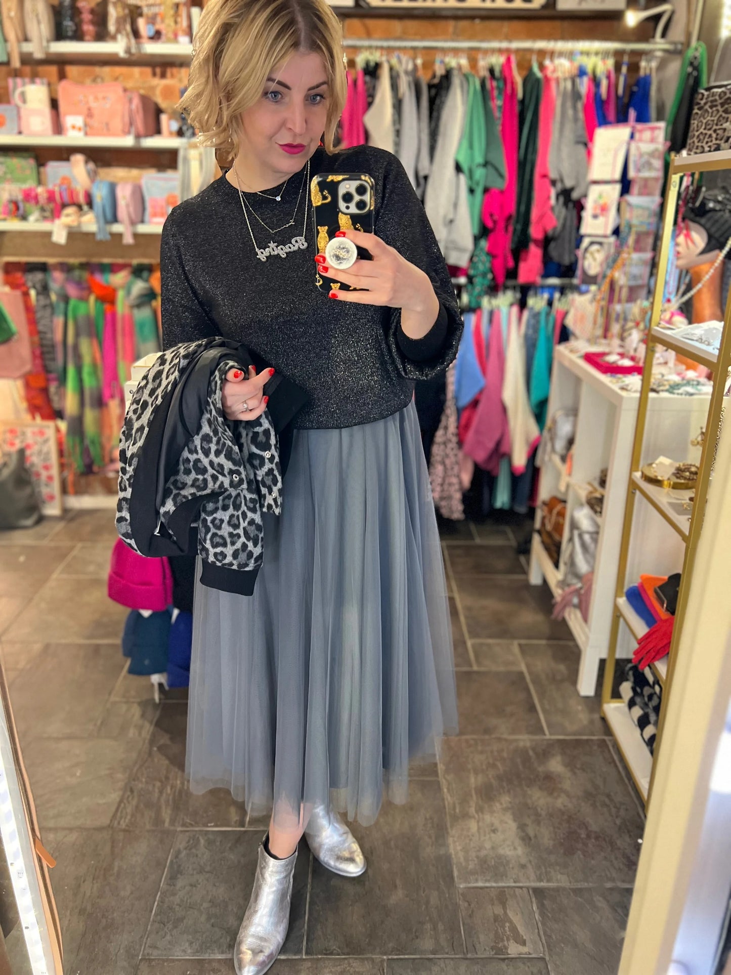 Woman taking a mirror selfie in a clothing store wearing a black sweater with a graphic design, gray tulle skirt, and silver boots.