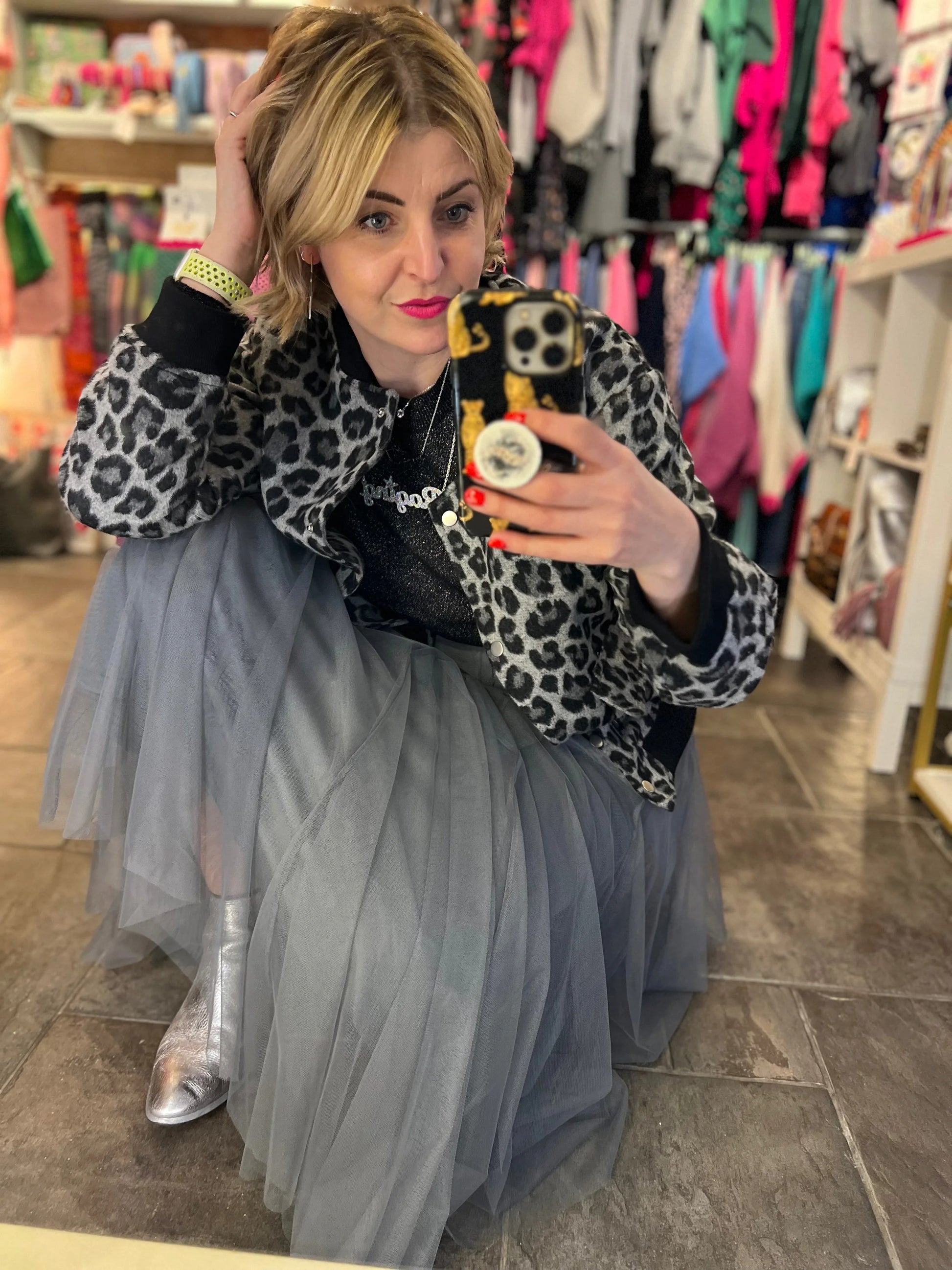 Woman taking a mirror selfie in a clothing store wearing a leopard print jacket and gray tulle skirt.