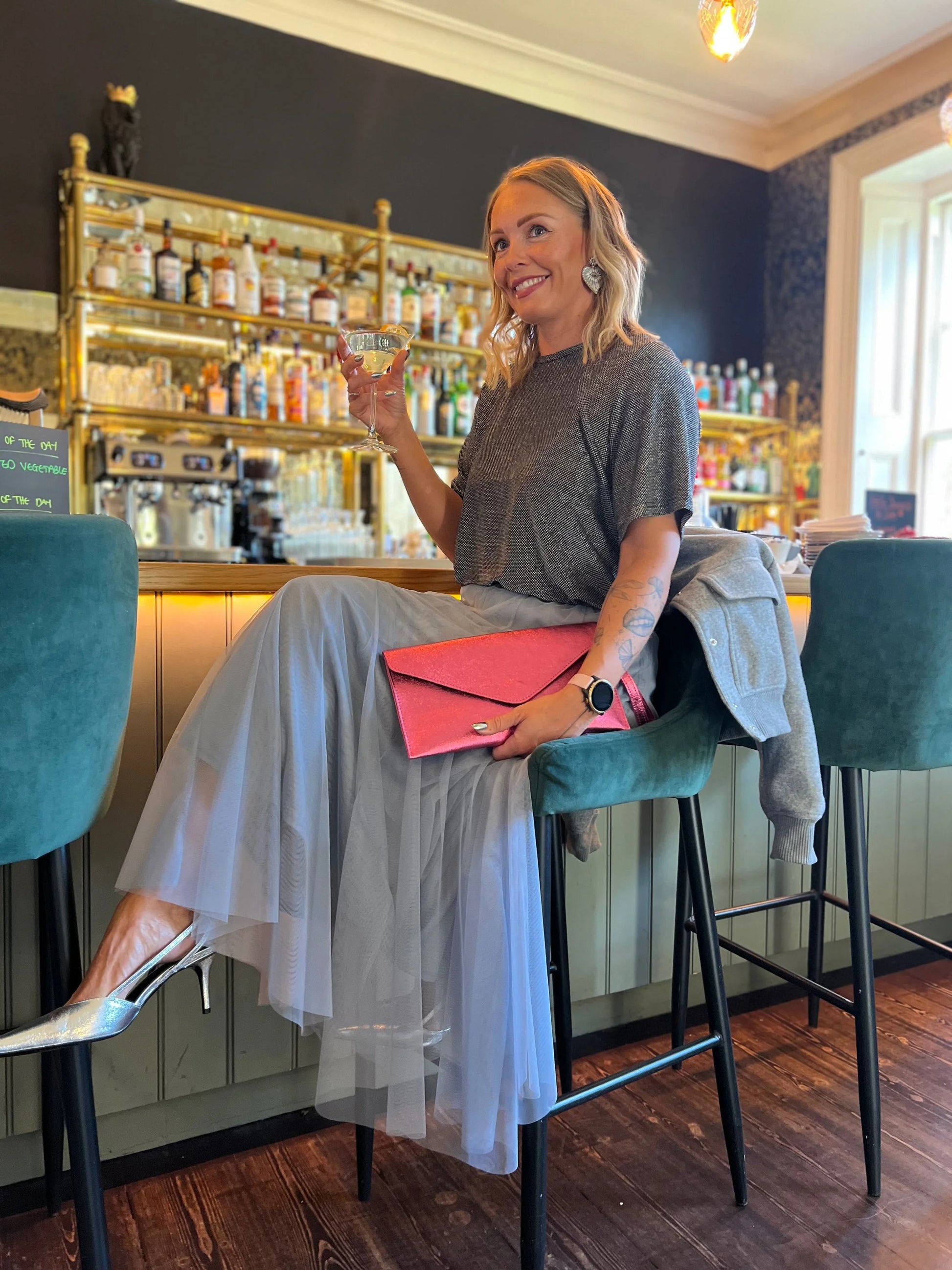 Woman sitting at a bar, holding a glass and a pink clutch, with a well-stocked bar in the background.