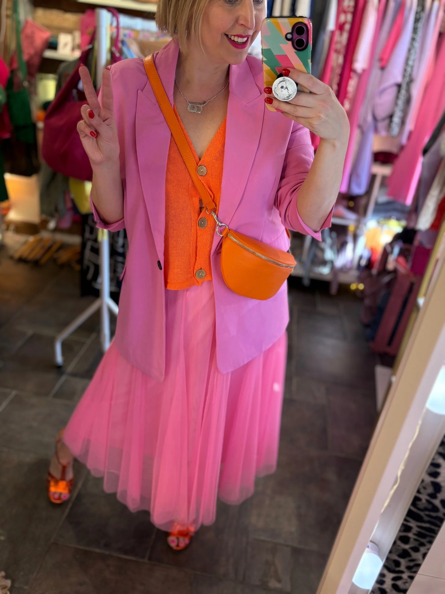 Person taking a mirror selfie wearing a pink outfit with an orange crossbody bag in a store.