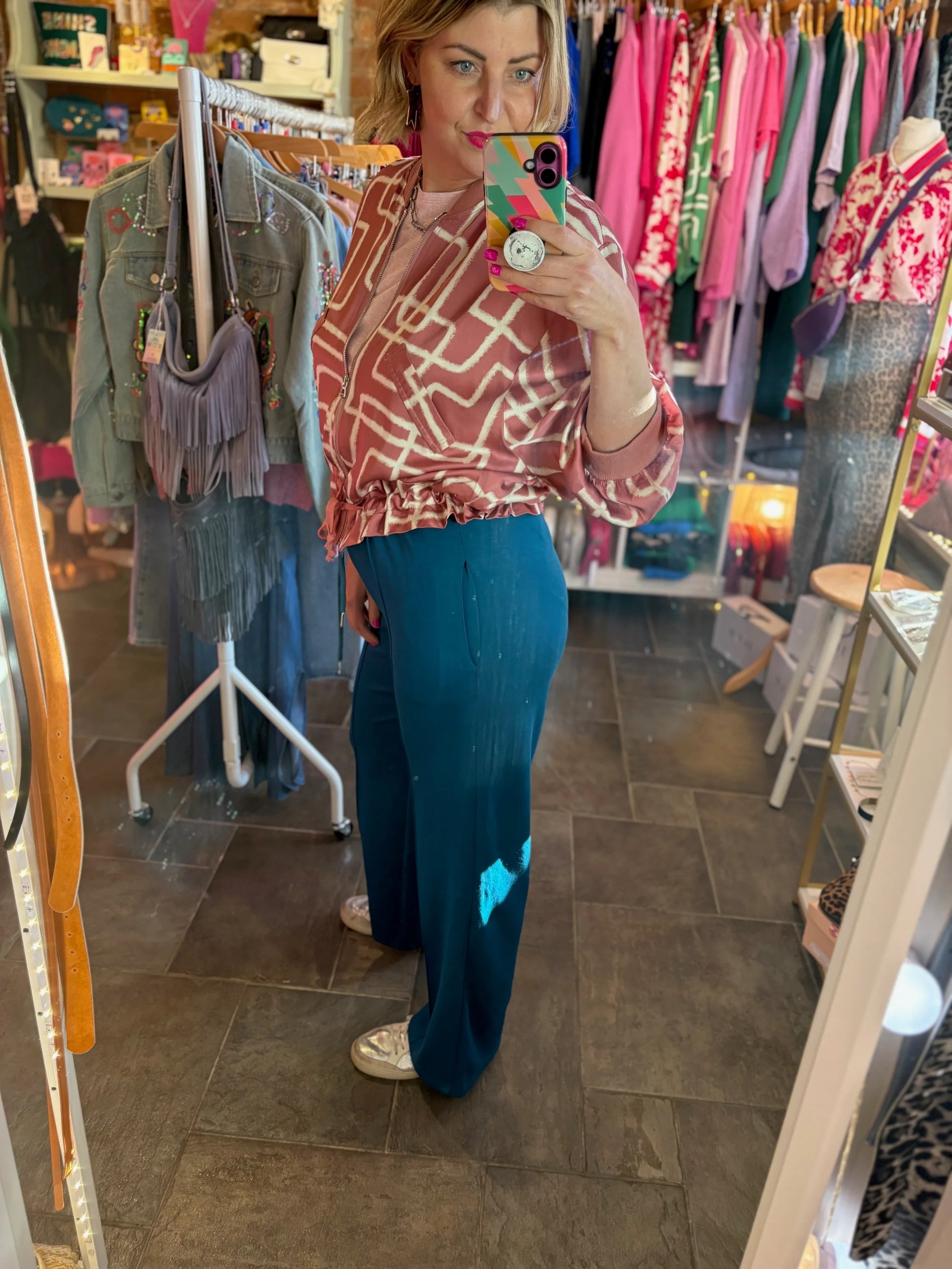 Woman taking a mirror selfie in a clothing store wearing a patterned top and blue pants.