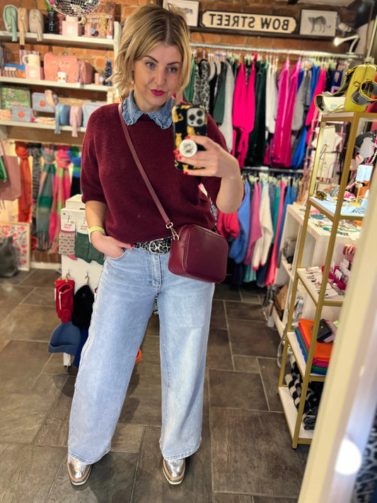 Woman taking a mirror selfie in a store wearing a burgundy sweater, light blue jeans, and a maroon handbag.