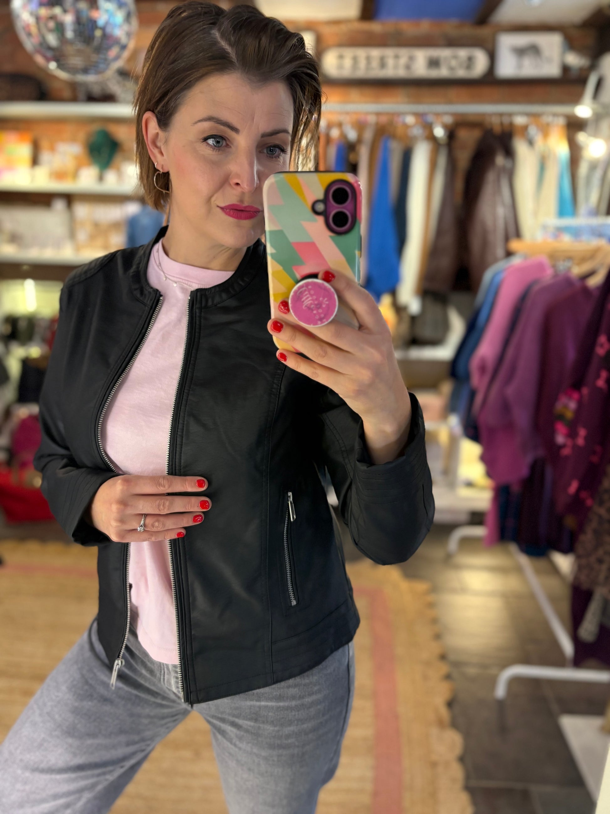 Woman taking a mirror selfie in a clothing store wearing a black biker jacket and grey trousers