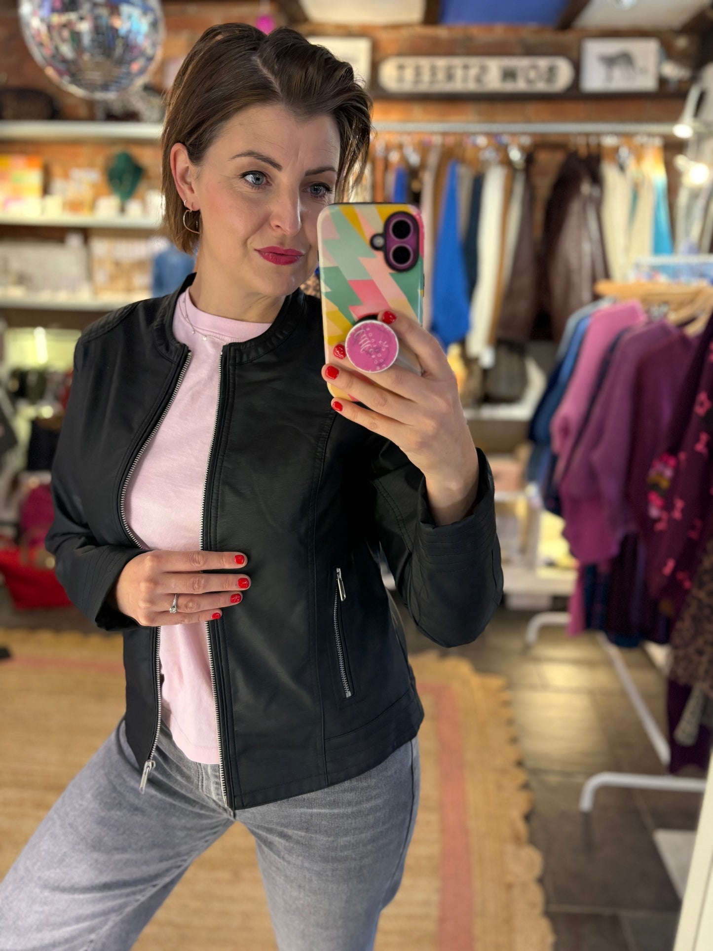 Woman taking a mirror selfie in a clothing store wearing a black jacket and pink tshirt and jeans