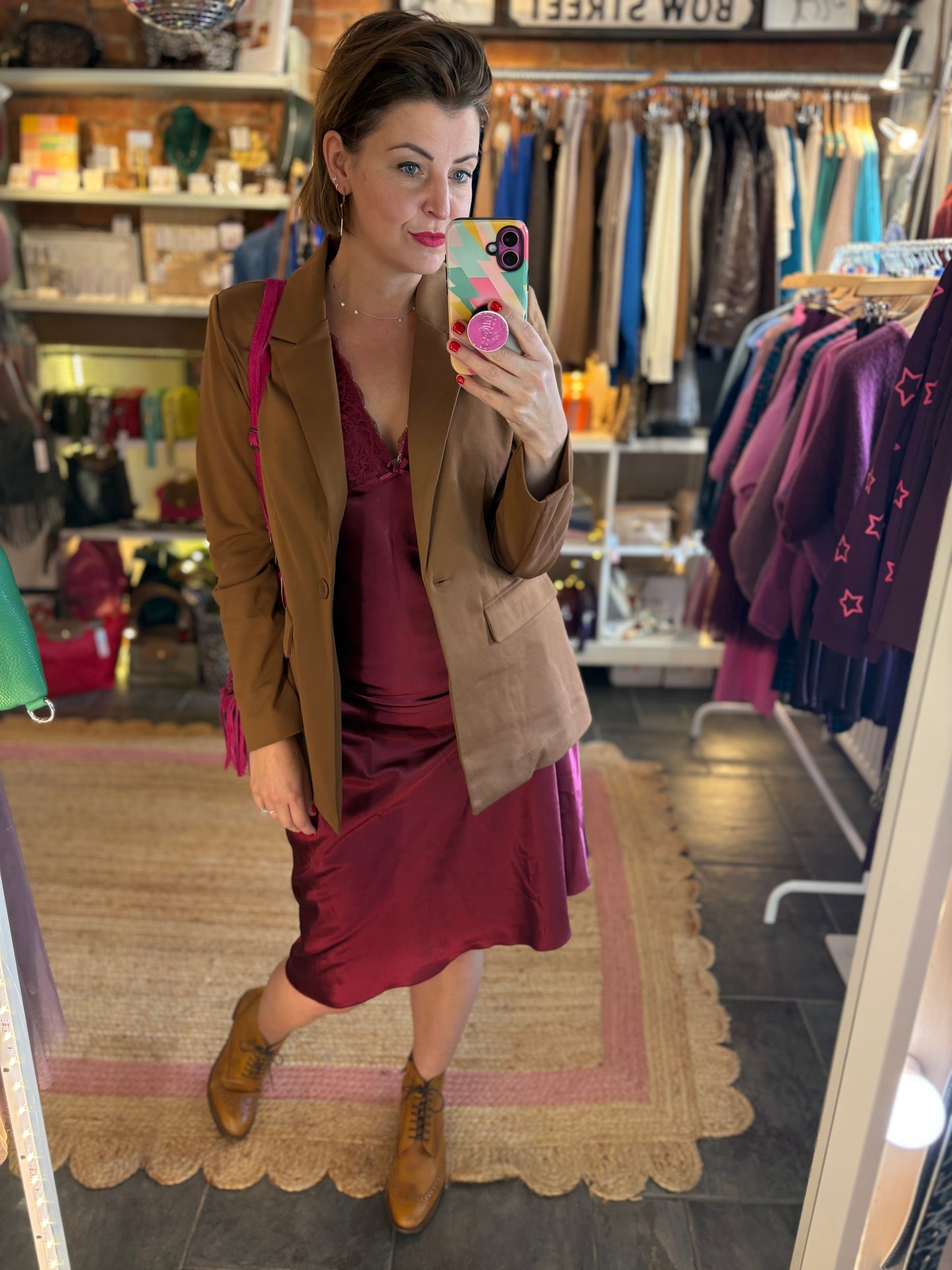 Woman taking a mirror selfie in a clothing store wearing a brown blazer and burgundy satin lace dress.