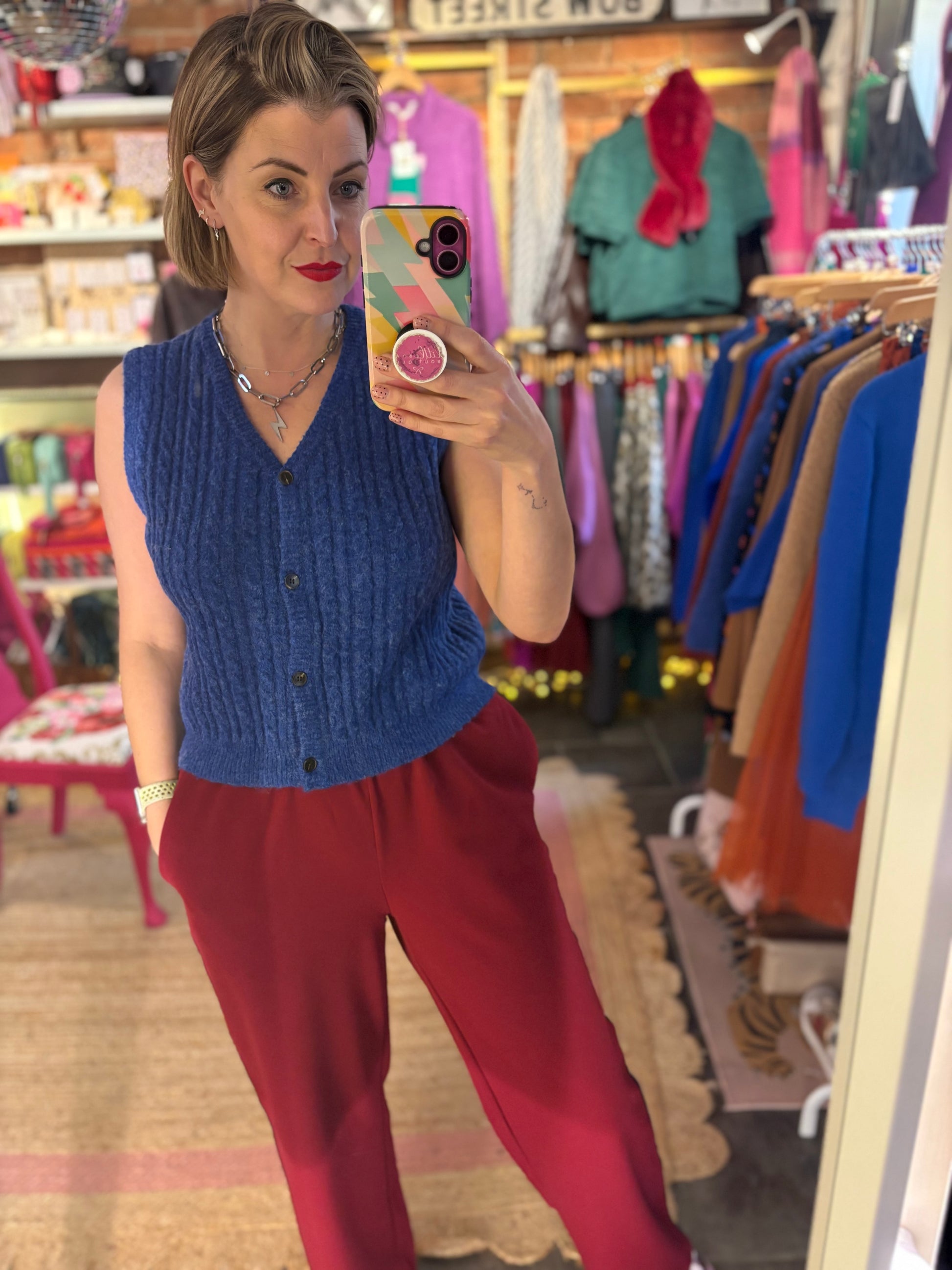 Woman taking a mirror selfie wearing a blue knitted vest and red trousers in a clothing store.