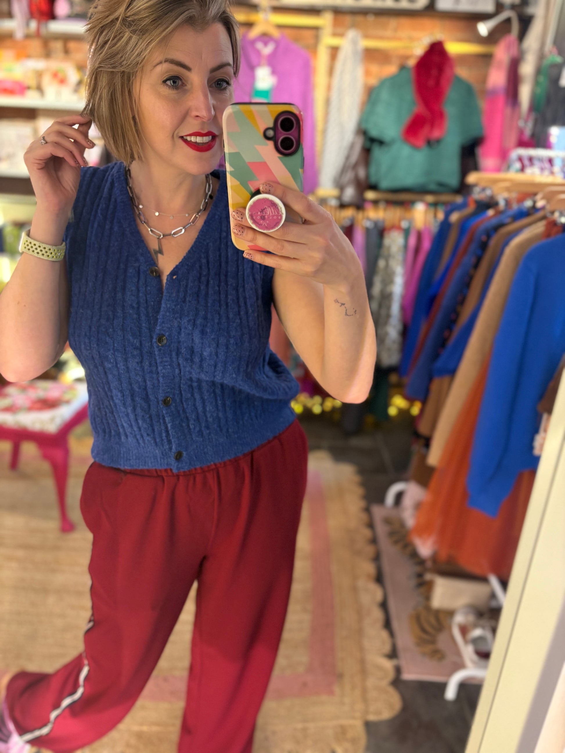 Woman taking a mirror selfie in a clothing store wearing a blue vest and red relaxed trousers
