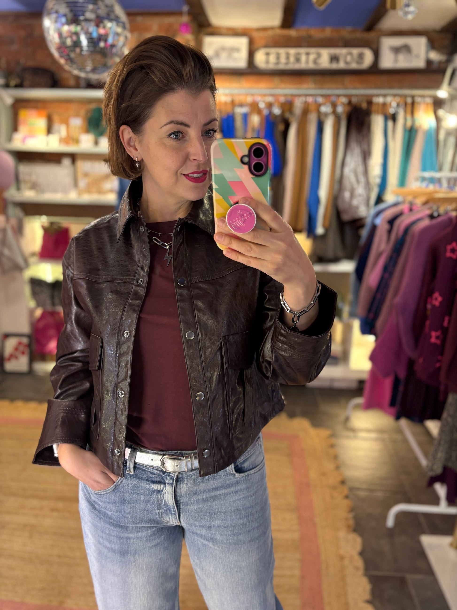 Woman taking a mirror selfie in a clothing store wearing a burgundy top and  brown leather jacket and blue jeans.