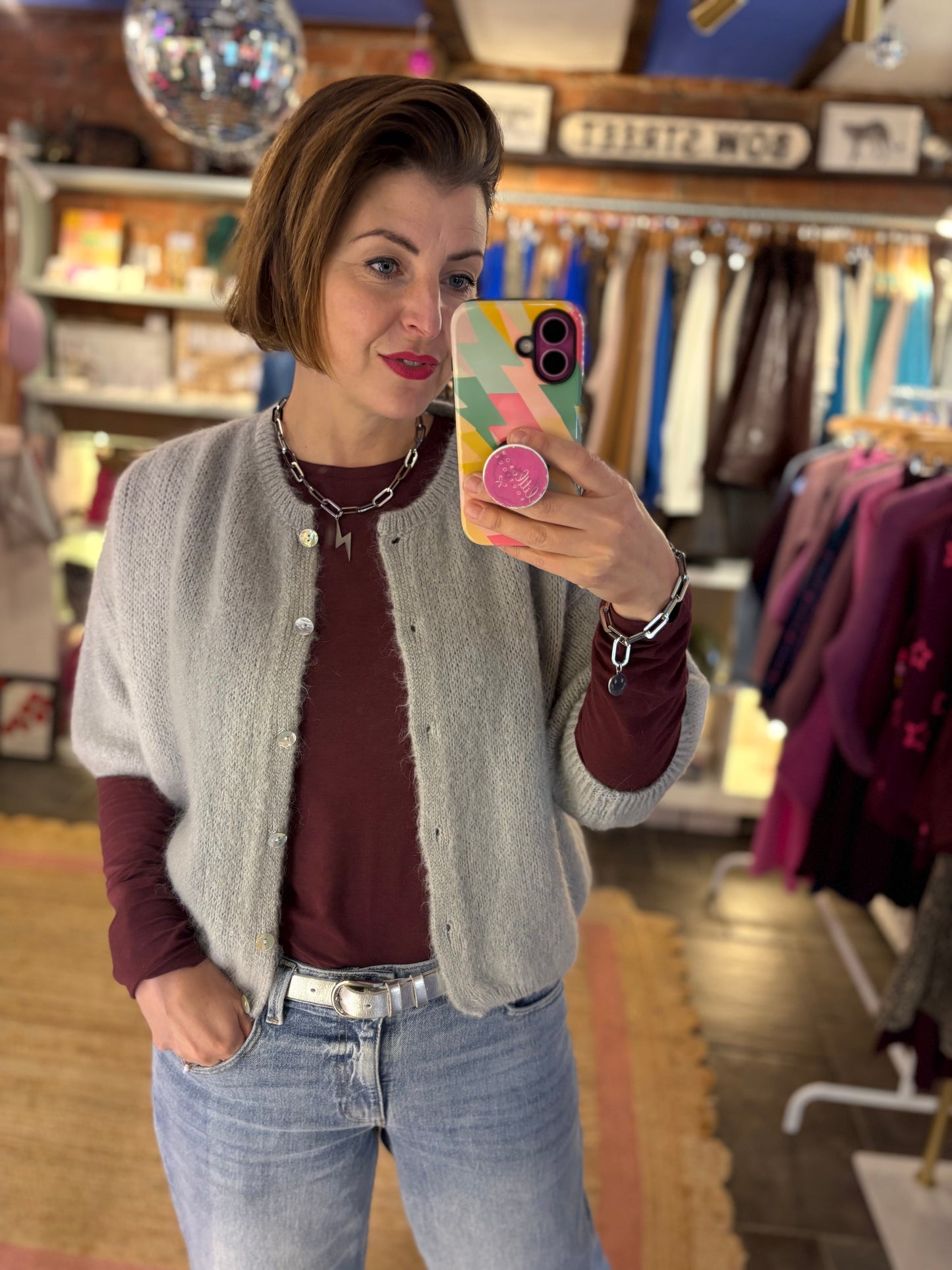 Woman taking a mirror selfie in a clothing store wearing a burgundy top layered with a light grey cardigan and blue jeans.