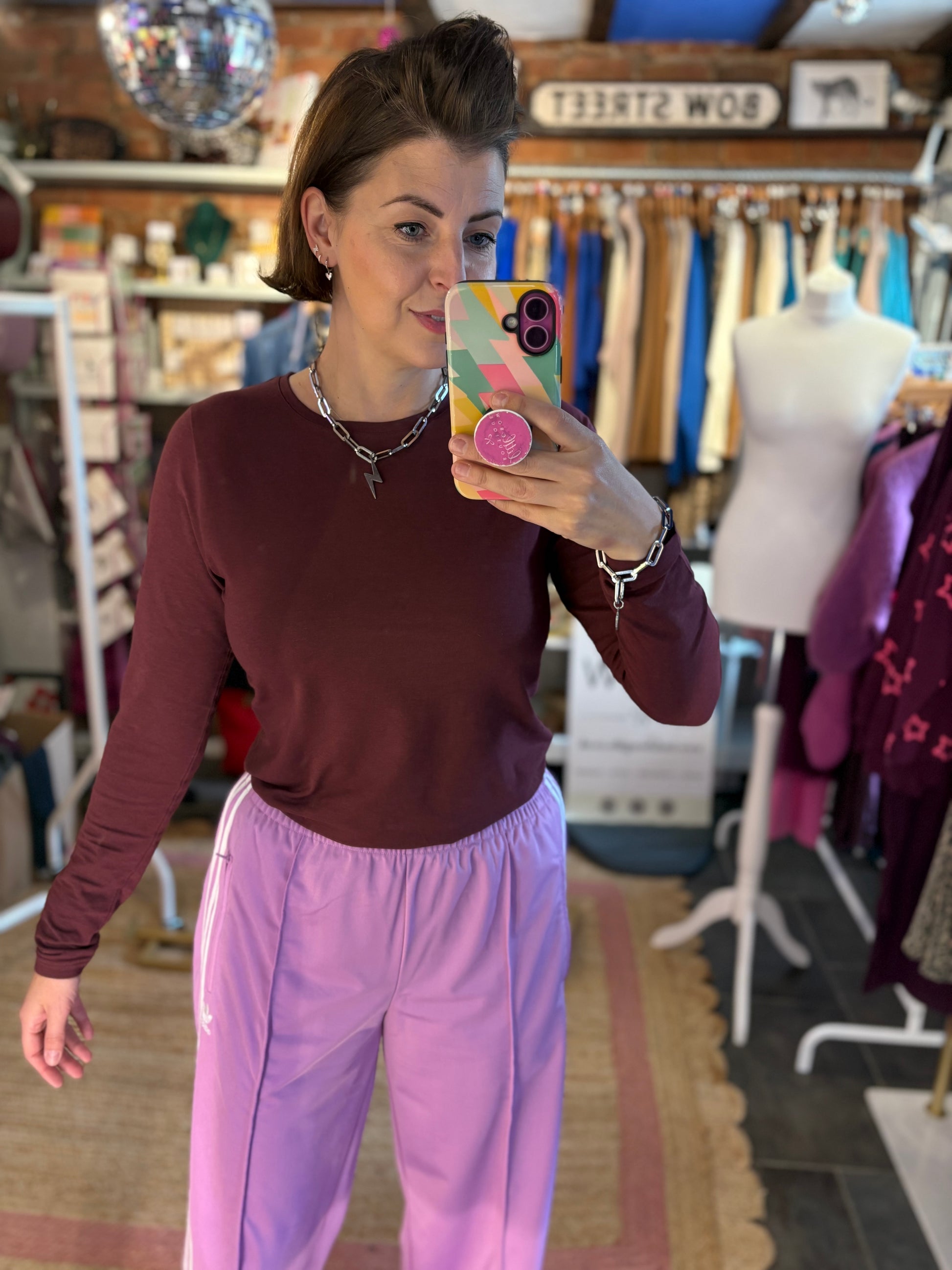 Woman taking a mirror selfie wearing a maroon long sleeve top  and lilac trousers in a clothing store.
