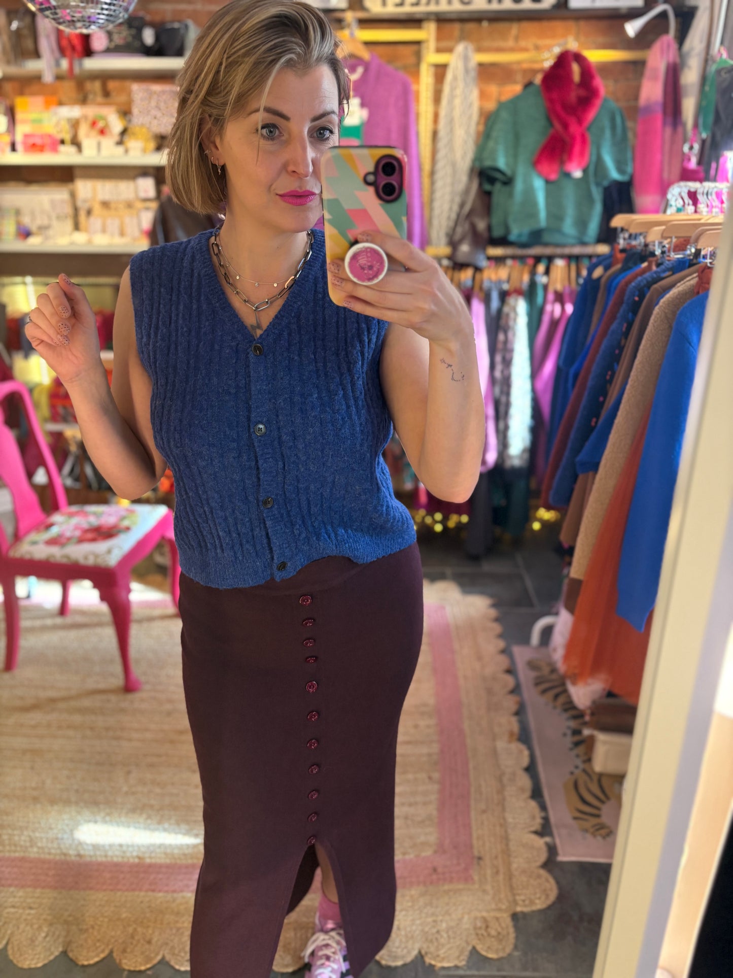 Woman taking a mirror selfie wearing a blue sleeveless top and burgundy/wine  skirt in a clothing store.