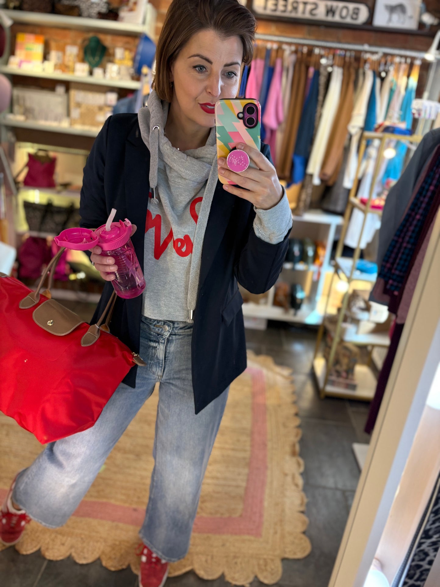 Woman taking a mirror selfie in a store, holding a pink drink and a red bag and blue jeans. Styled for a Sports Luxe Look.