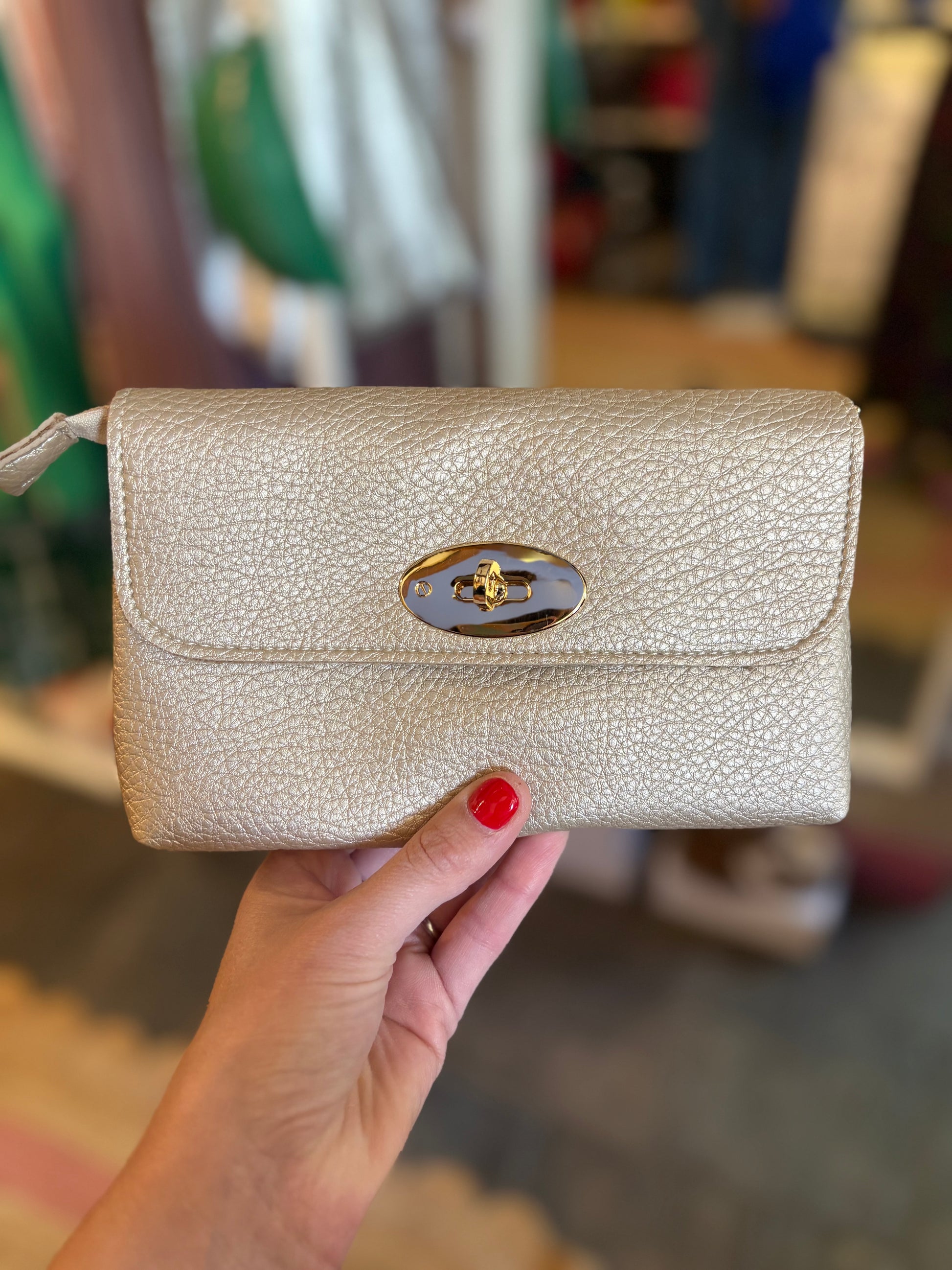 Hand holding a beige metallic  textured handbag with a gold clasp in an indoor setting.