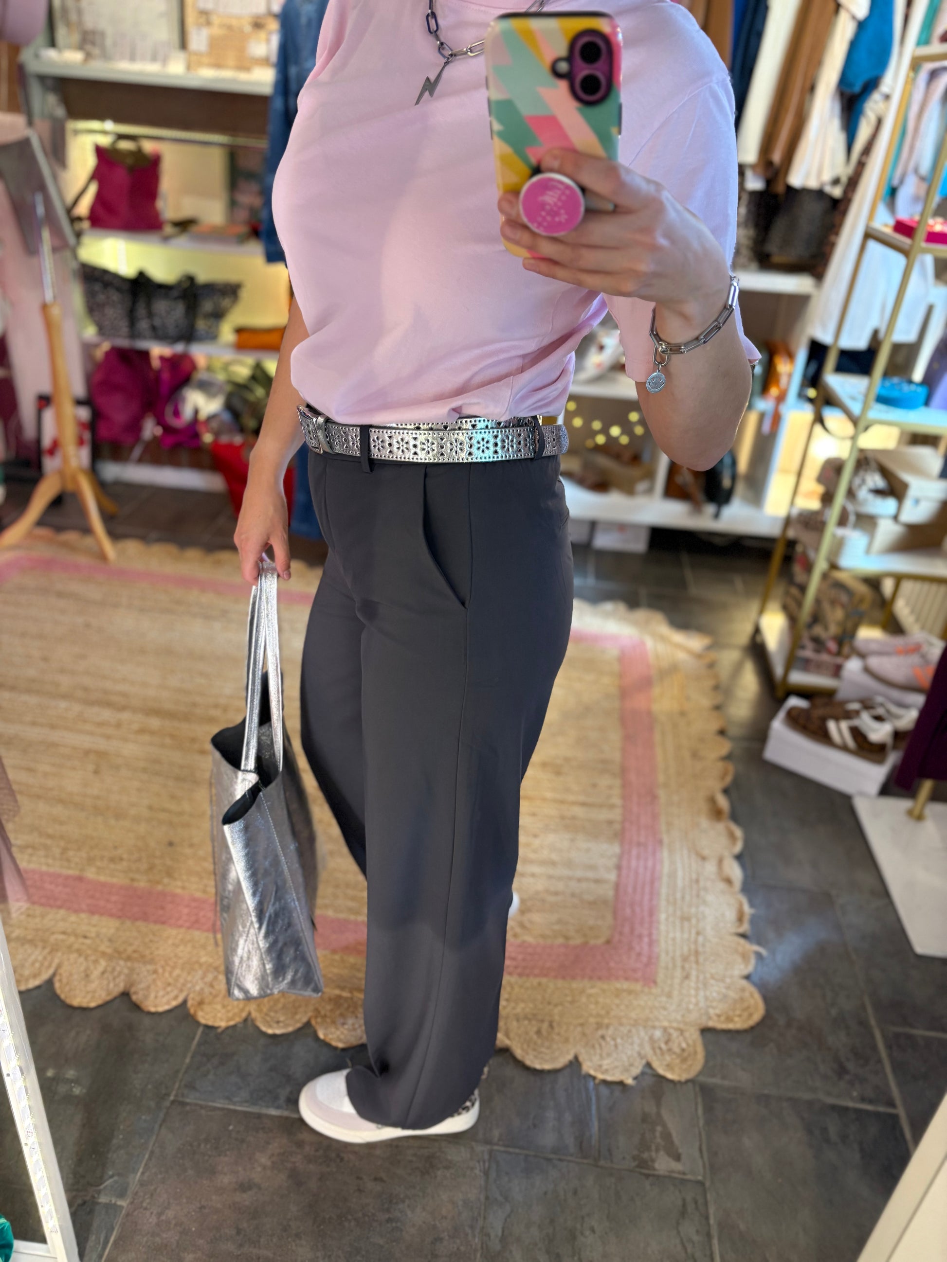 Person taking a mirror selfie wearing a pink t-shirt, grey trousers, and white trainers in a store.