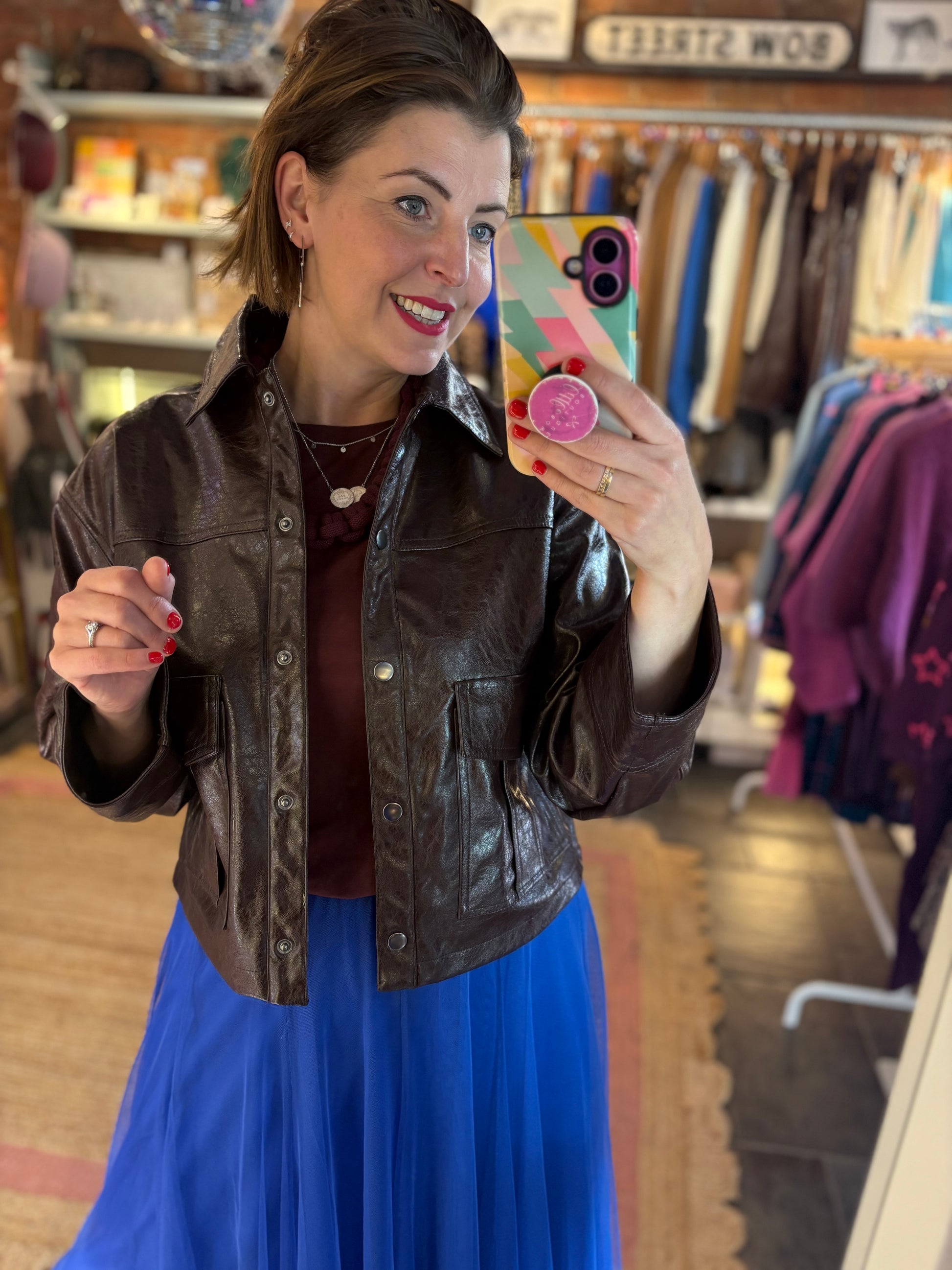 Woman taking a mirror selfie wearing a brown leather jacket and blue skirt in a store.