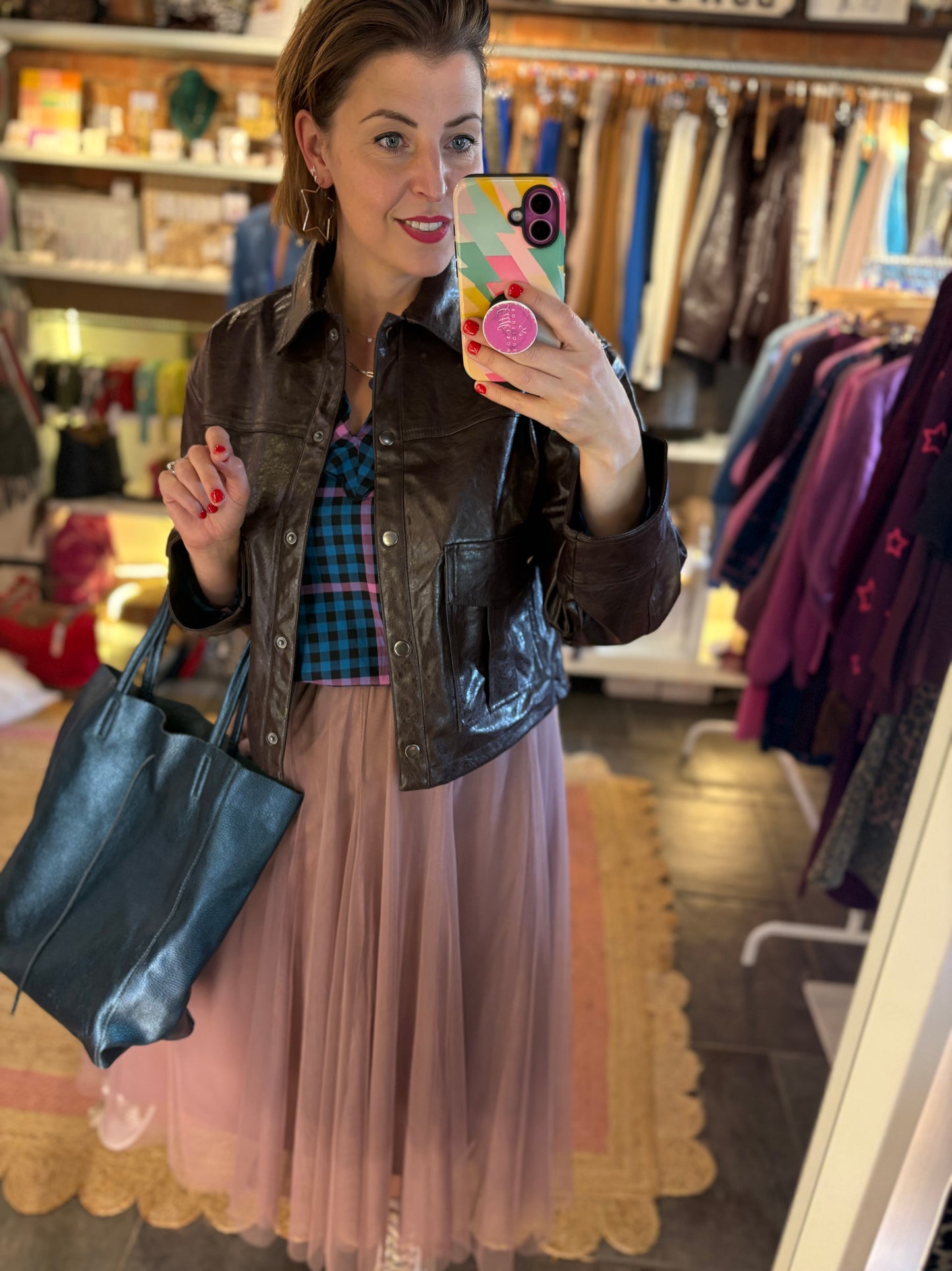 Woman taking a mirror selfie in a clothing store wearing a brown jacket, plaid shirt, and pink tulle skirt.