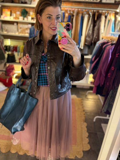 Woman taking a mirror selfie in a clothing store wearing a brown jacket, plaid shirt, and pink tulle skirt.