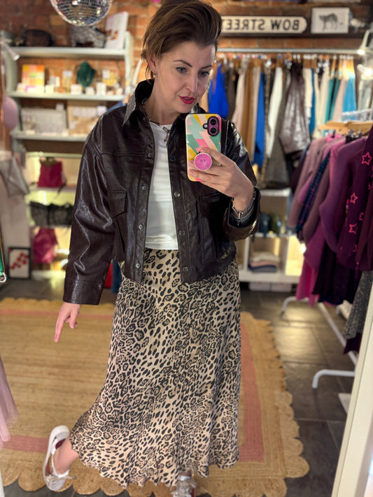 Woman taking a mirror selfie wearing a black leather jacket and leopard print skirt in a store.