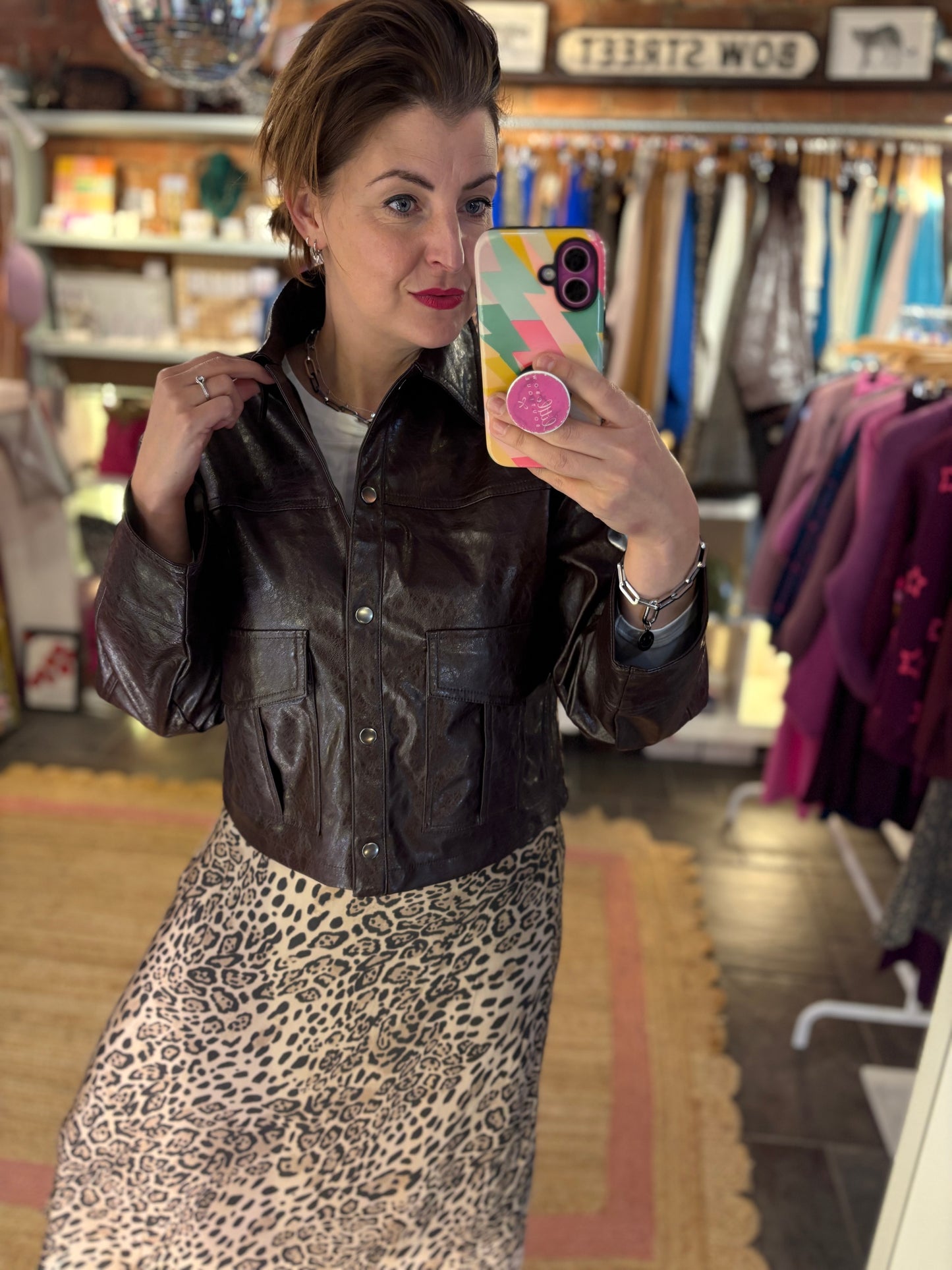 Woman taking a mirror selfie wearing a brown leather jacket and leopard print skirt in a store.