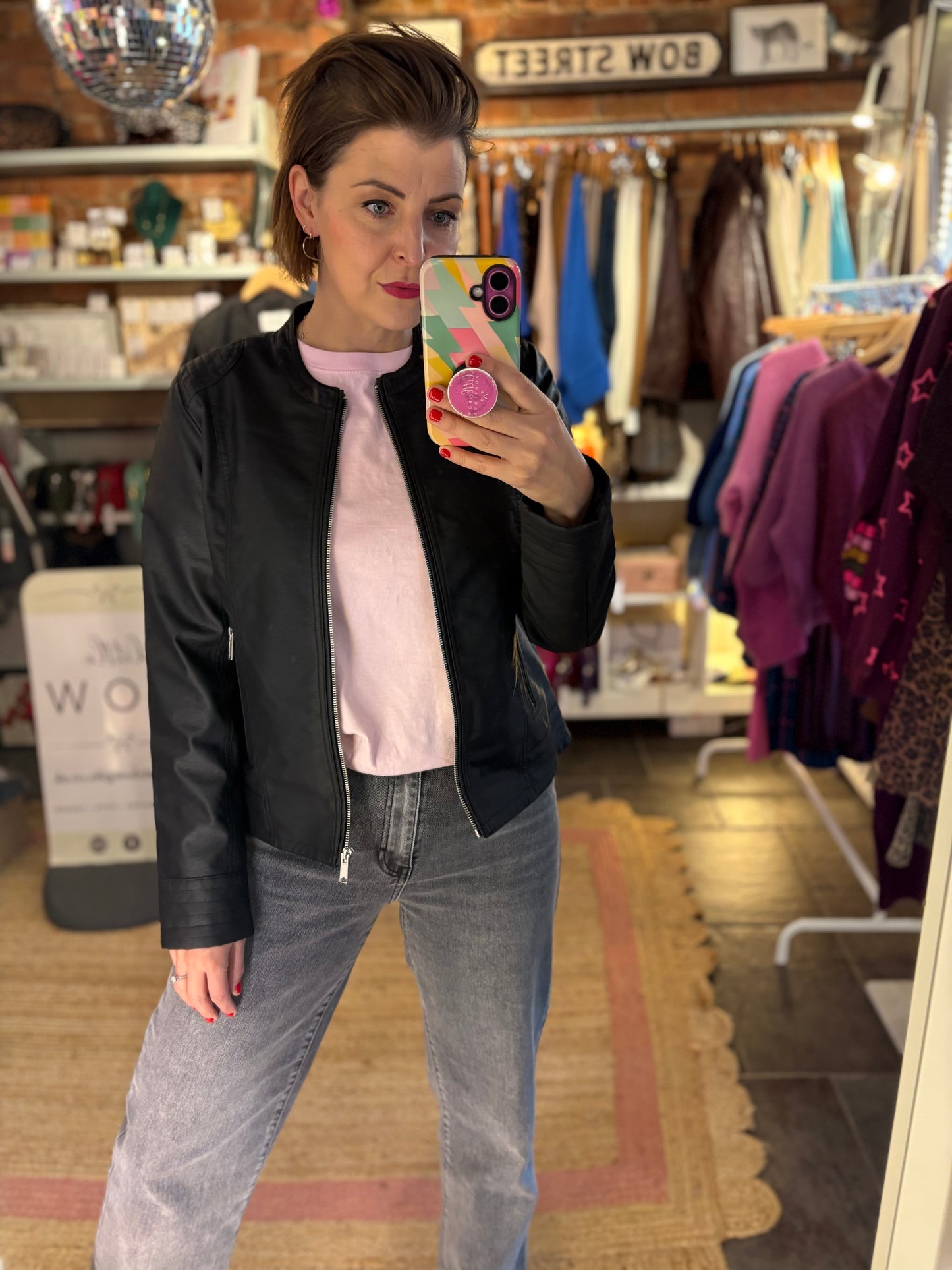 Woman taking a mirror selfie in a clothing store wearing a black jacket, pink tshirt, and grey jeans.