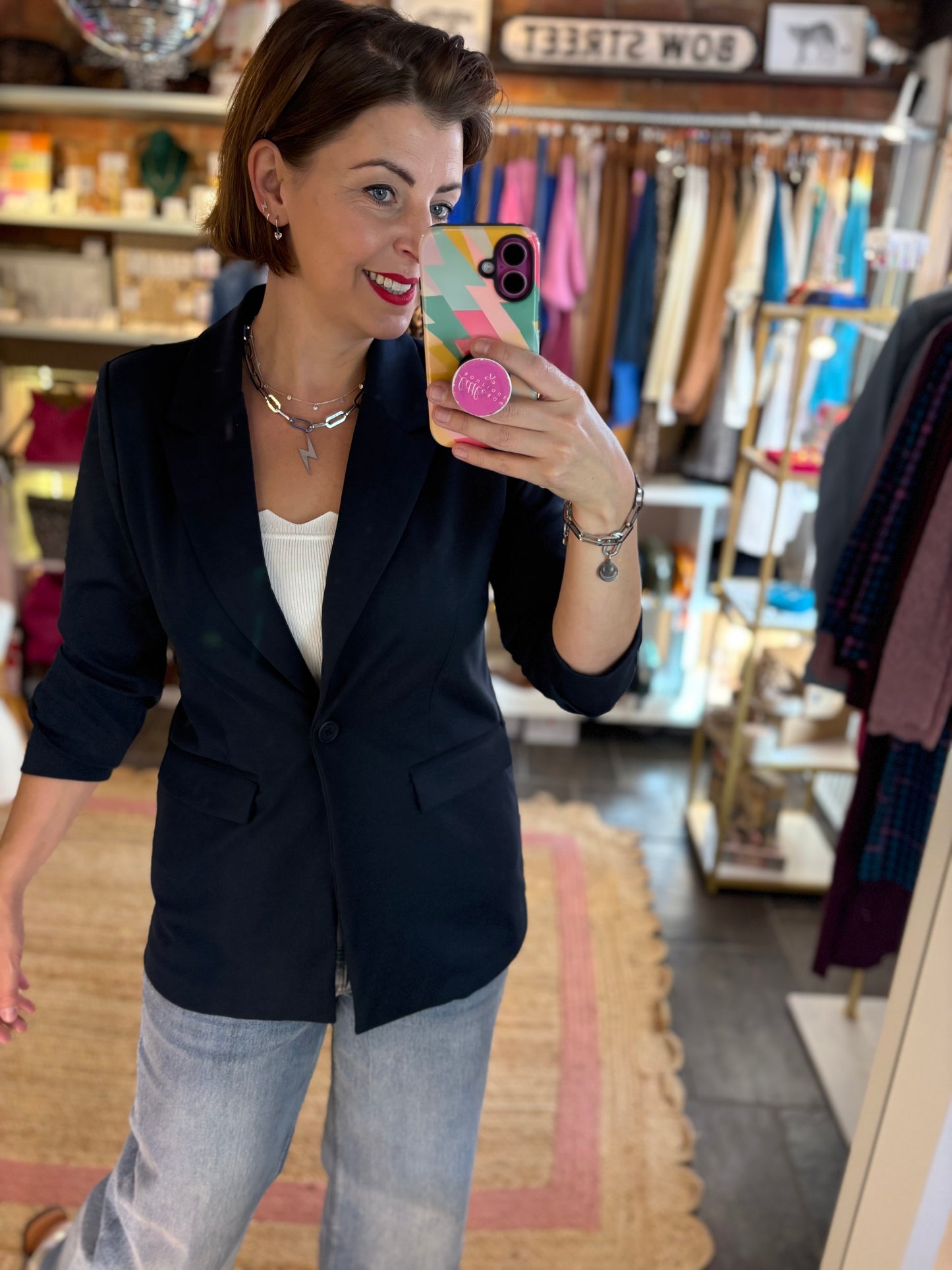 Woman taking a mirror selfie wearing a classic navy blazer and light blue jeans in a clothing store.